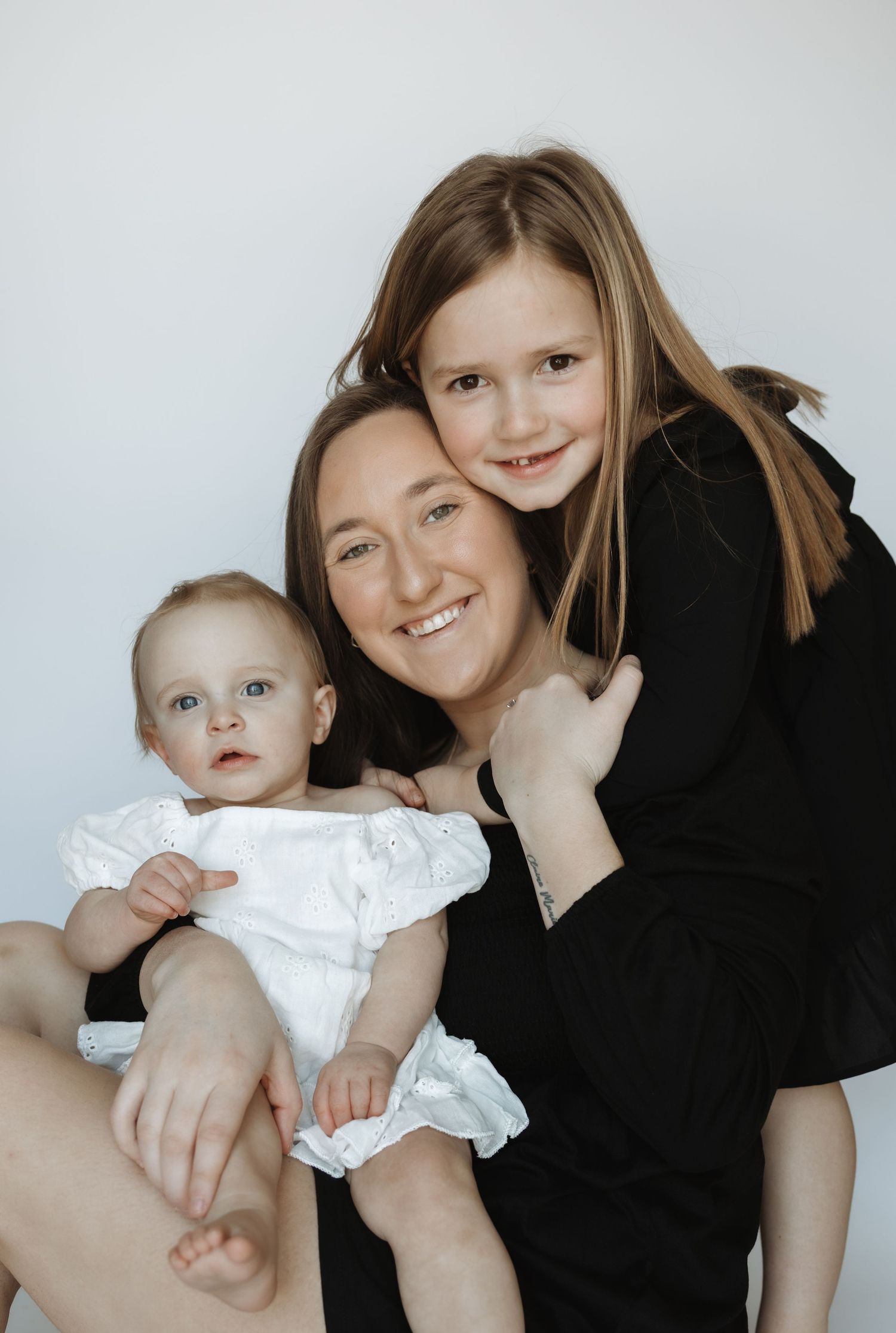Smiling mother in black dress holding baby in white outfit with older daughter hugging her from behind.