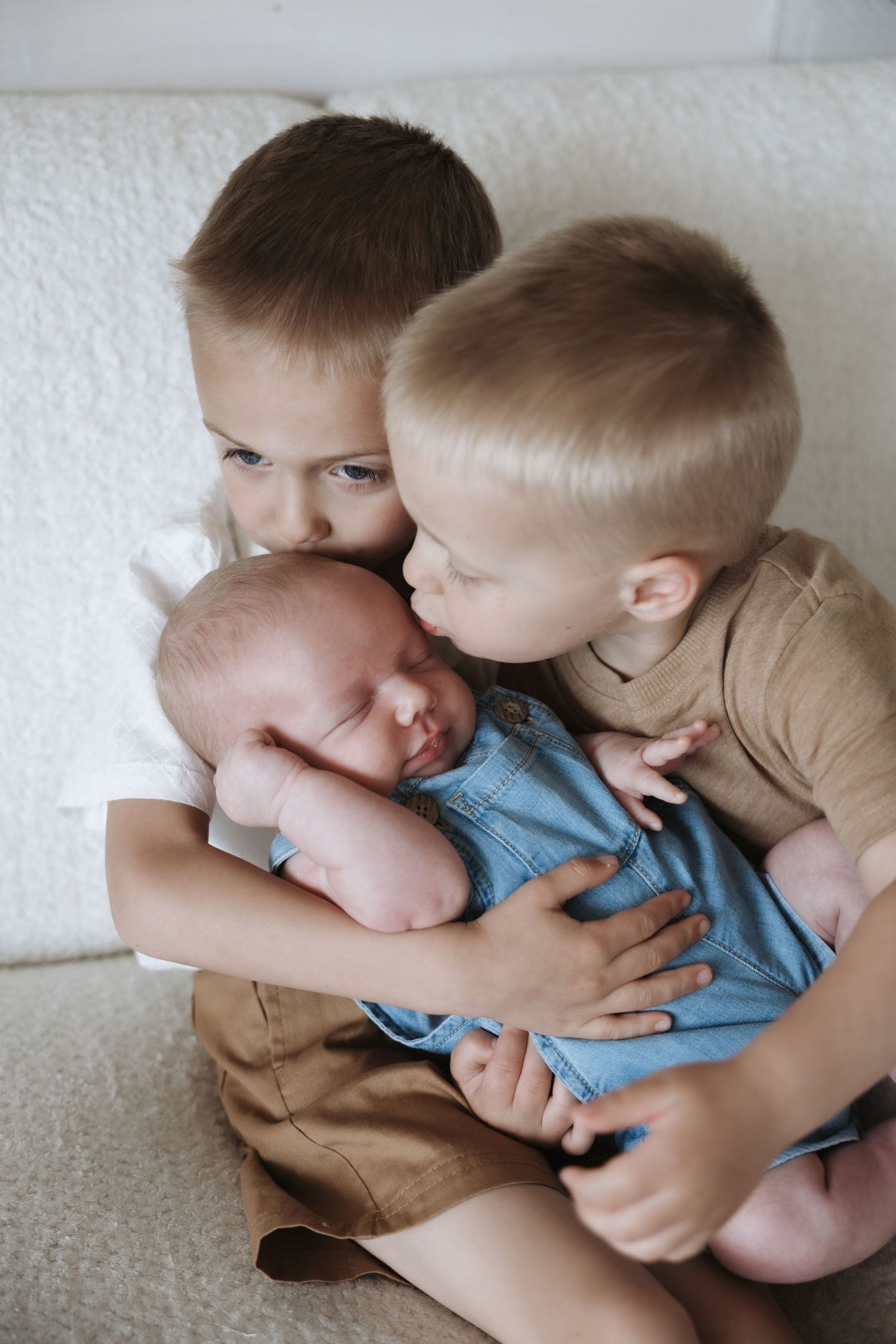 Two young boys lovingly hold and kiss their sleeping newborn baby sibling on a white couch.