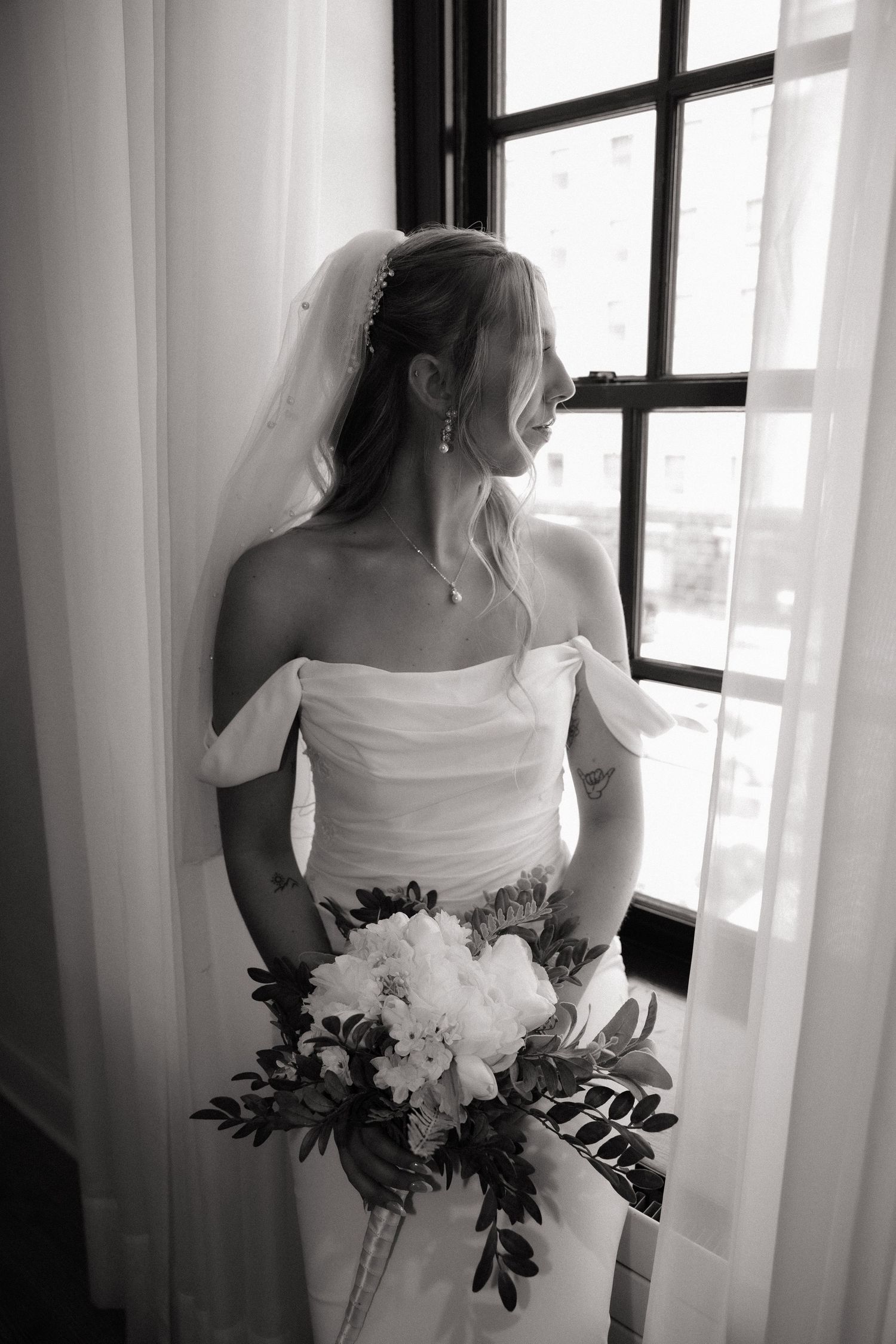 Bride in off-shoulder gown and veil holds white peony bouquet while gazing out window in black and white photo.