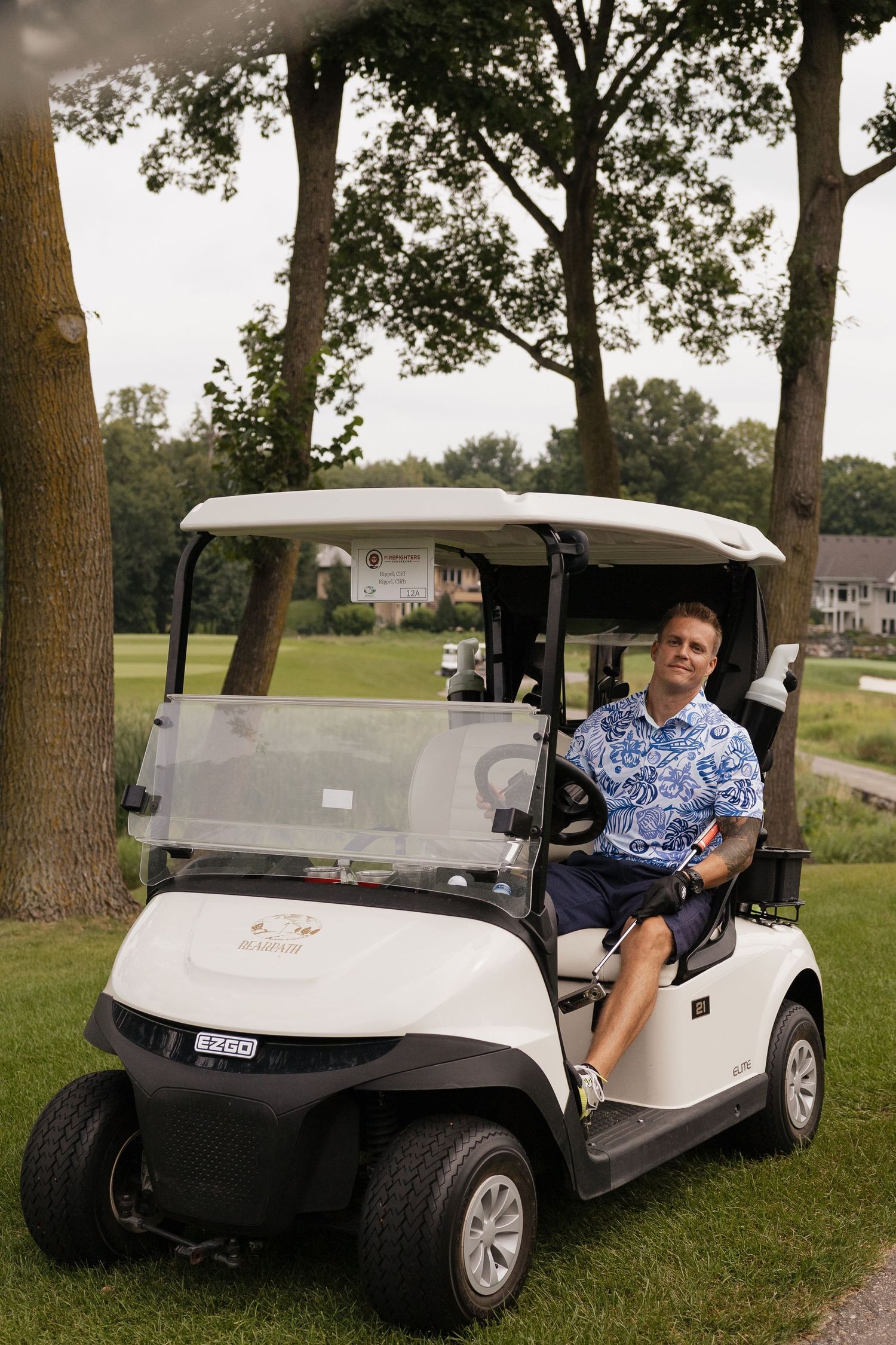 Man in blue floral shirt sitting in a white E-Z-GO golf cart on a lush green golf course.