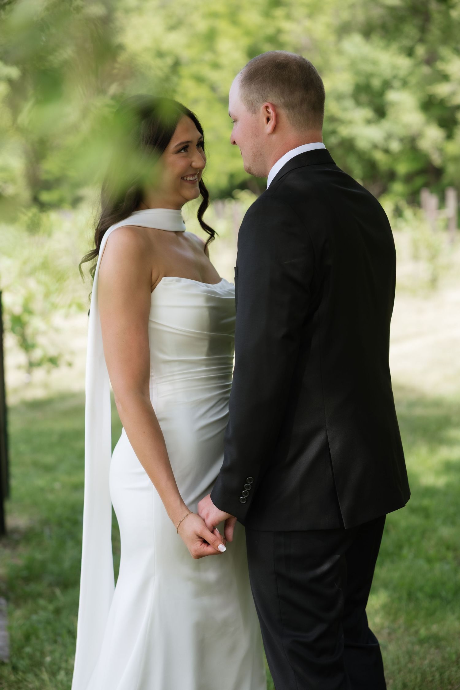 Bride and groom hold hands and share an intimate moment outdoors, surrounded by lush green trees.