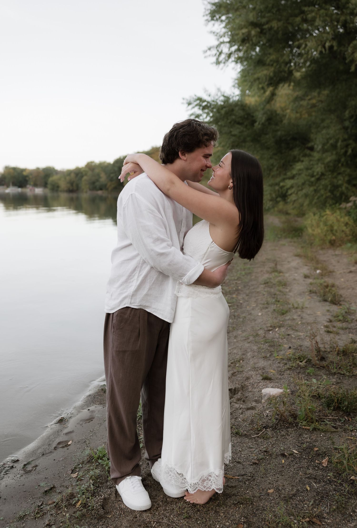 Couple embracing romantically by a serene lakeside, woman in white dress and man in white shirt and brown pants.