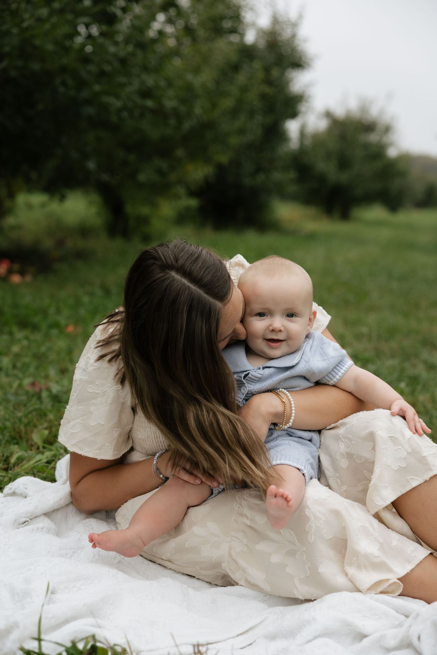 Mother kissing her smiling baby on a white blanket in a lush green garden during an outdoor family photo session.