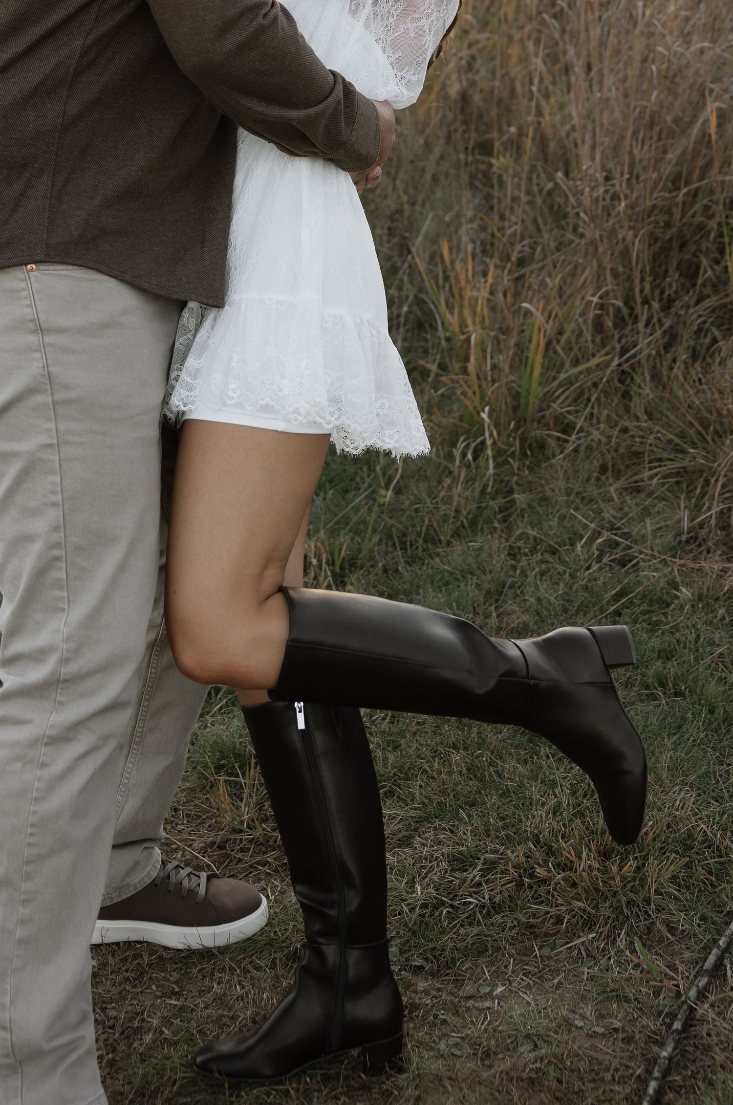 Couple embracing outdoors, woman in white dress and tall black riding boots, man in khaki pants and sneakers.