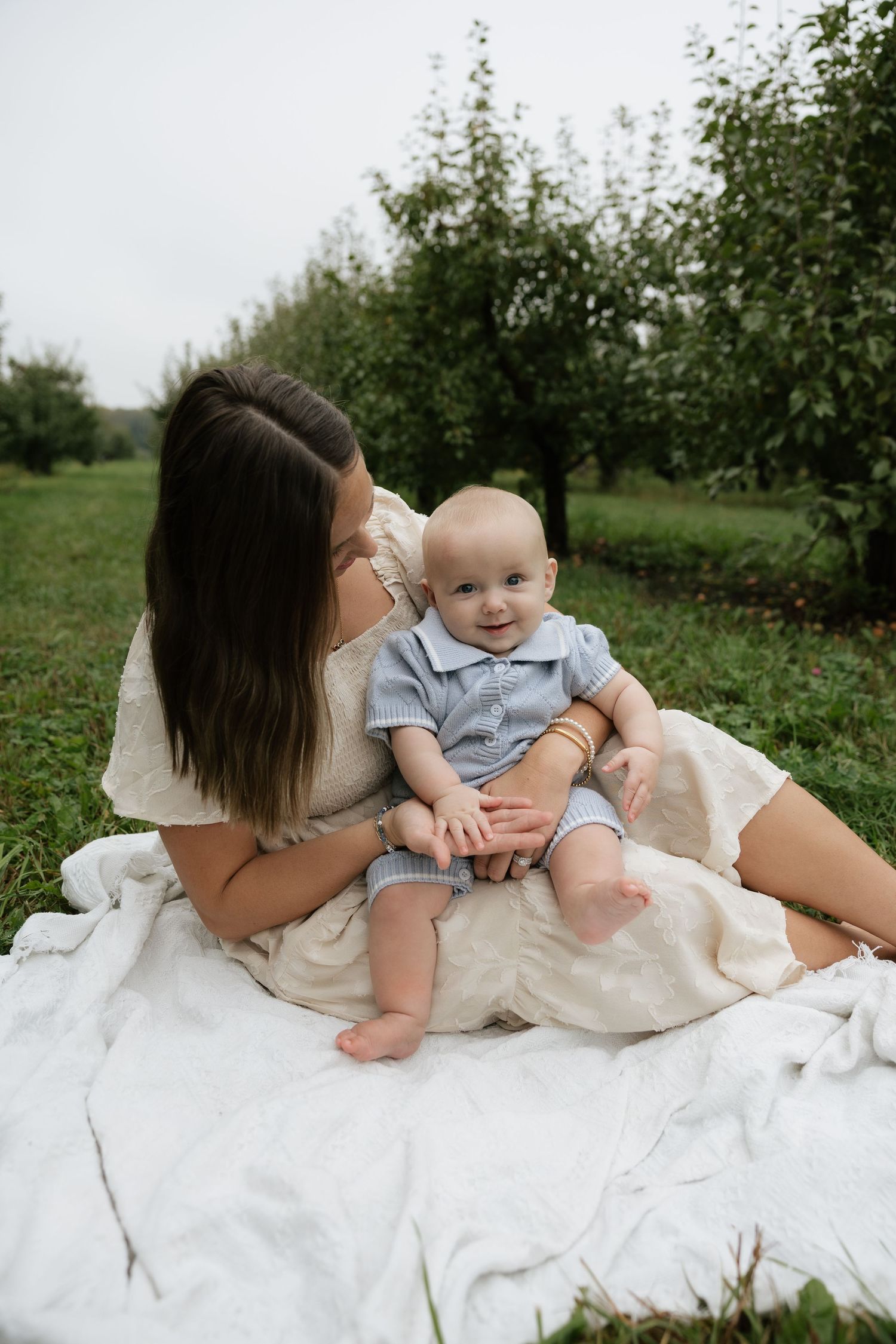 A young mother sits on a white blanket outdoors, lovingly holding her smiling baby in a lush green orchard.