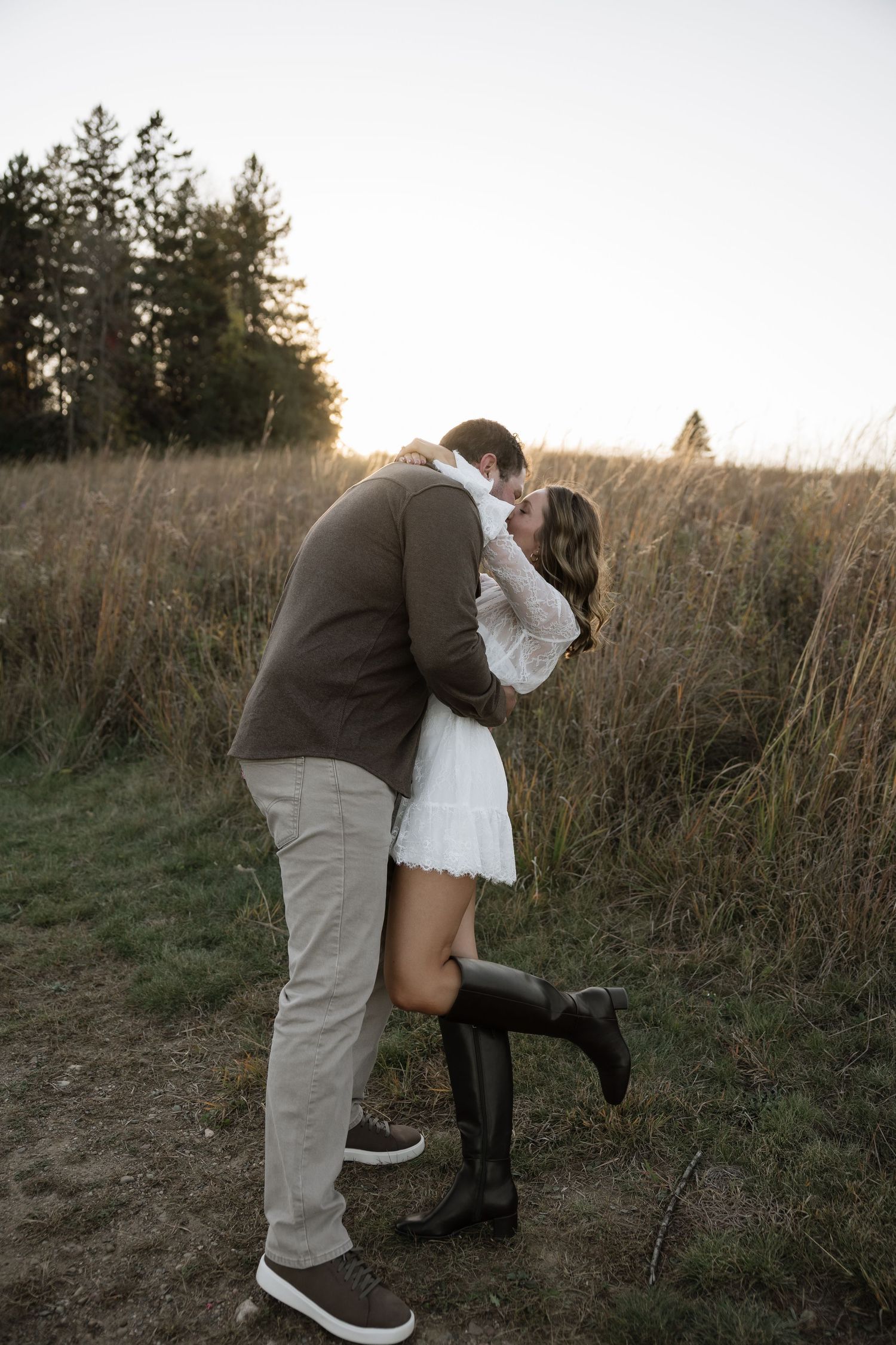 Couple sharing a romantic kiss in a golden field at sunset, woman in white dress and tall boots, man in brown jacket.