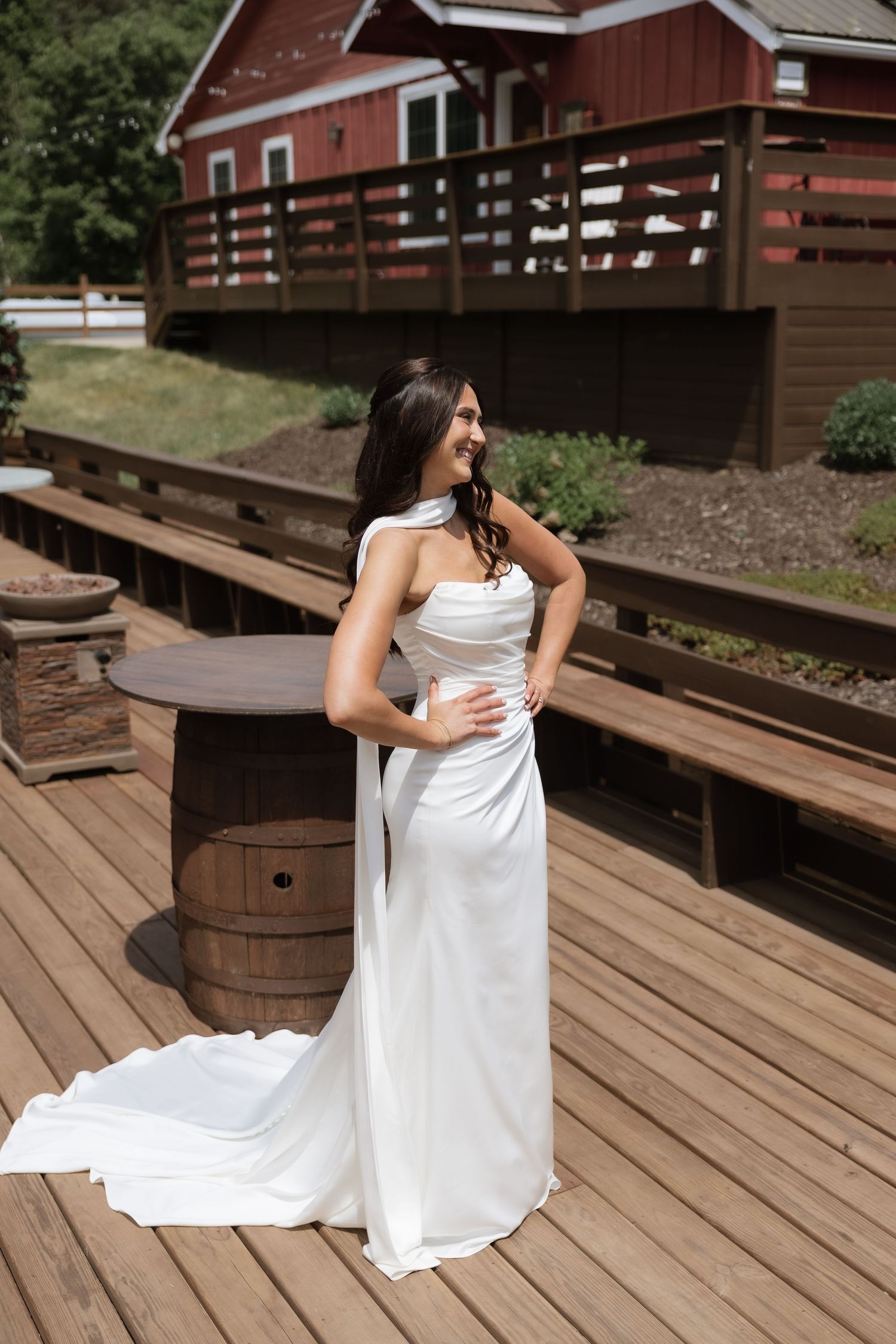 Elegant bride in sleek white gown with cape detail poses on wooden deck outside a red barn venue.