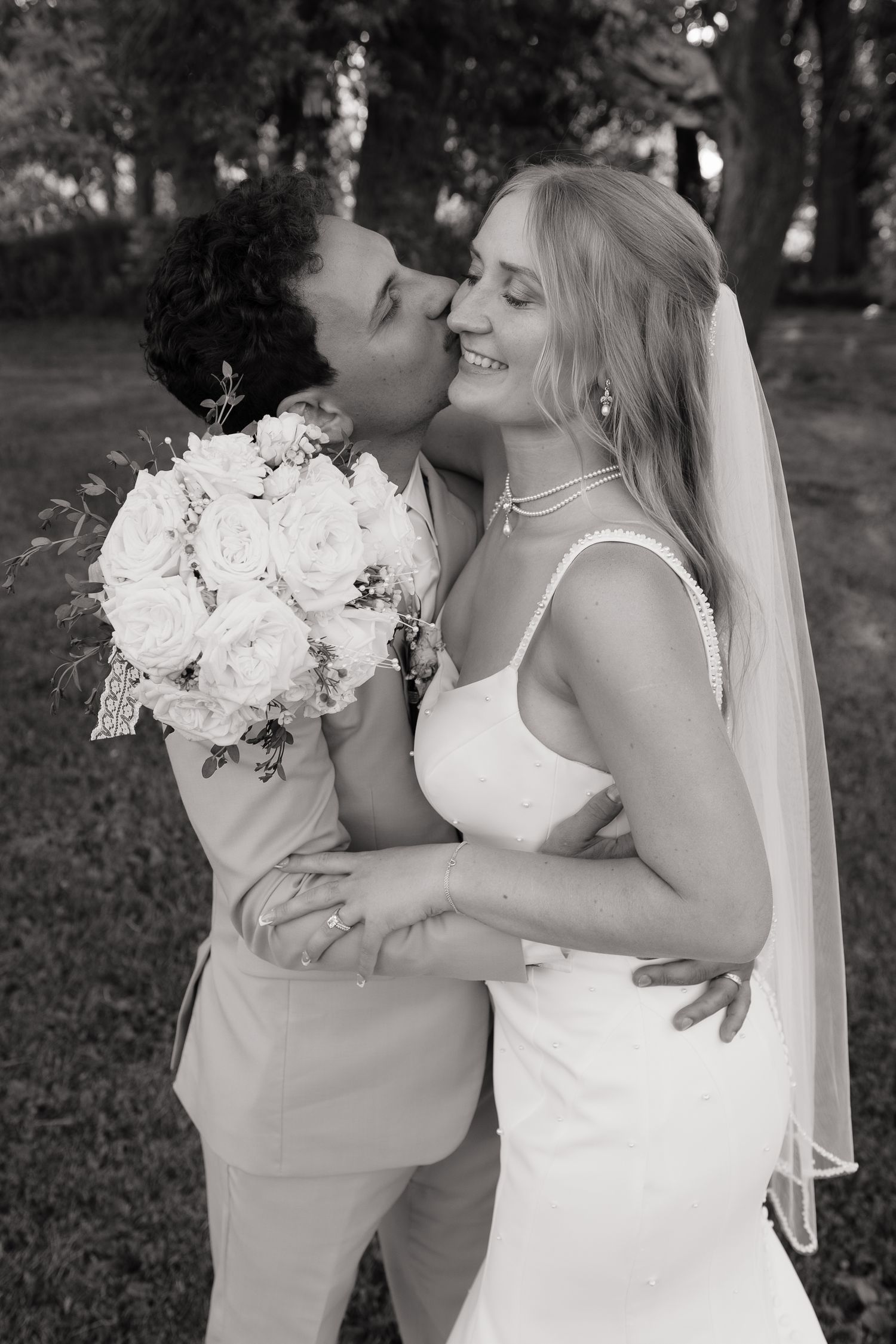 Black and white photo of groom kissing bride on cheek while she smiles holding white rose bouquet outdoors.