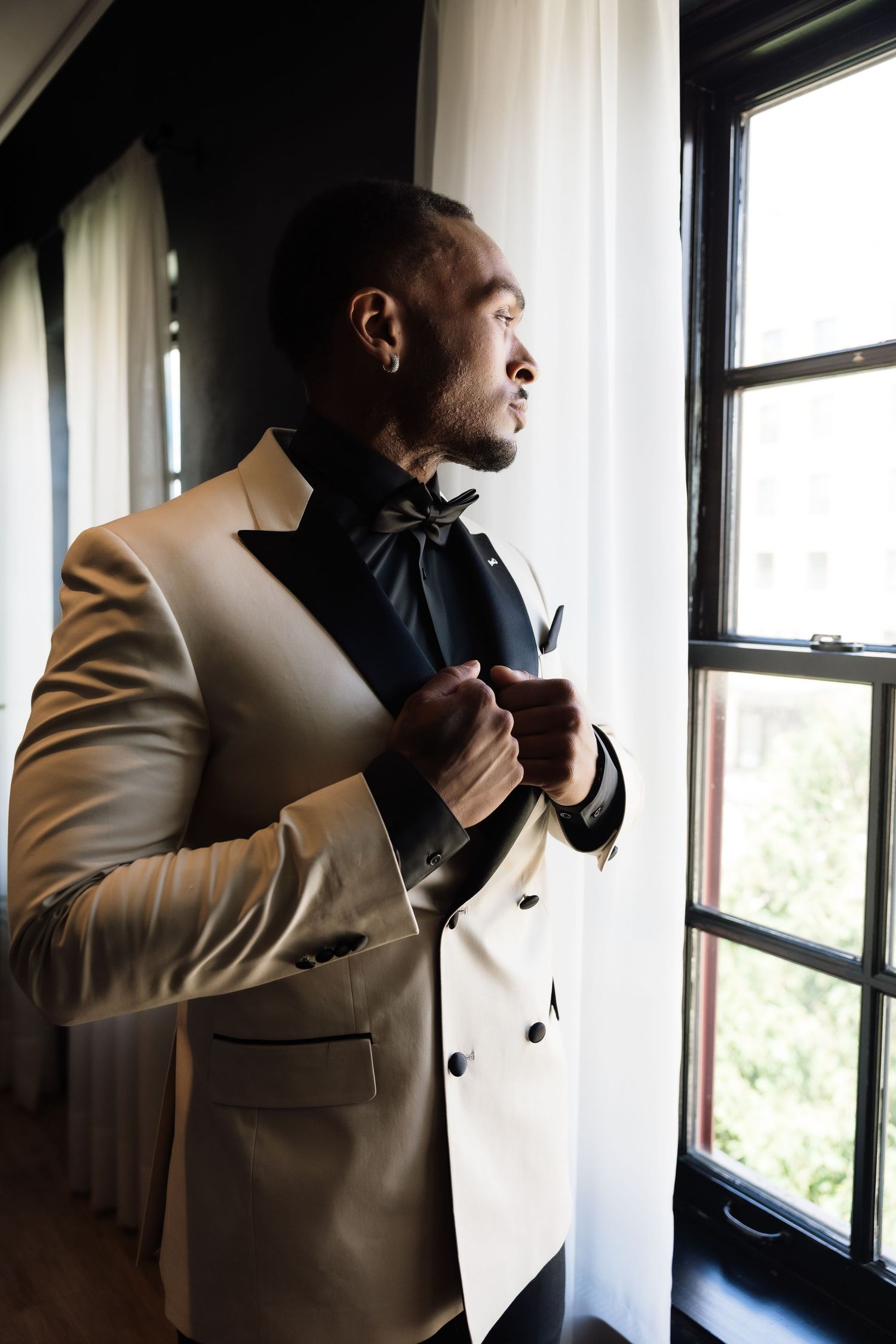 Elegant groom in white tuxedo jacket and black bow tie adjusting suit by window in natural light.