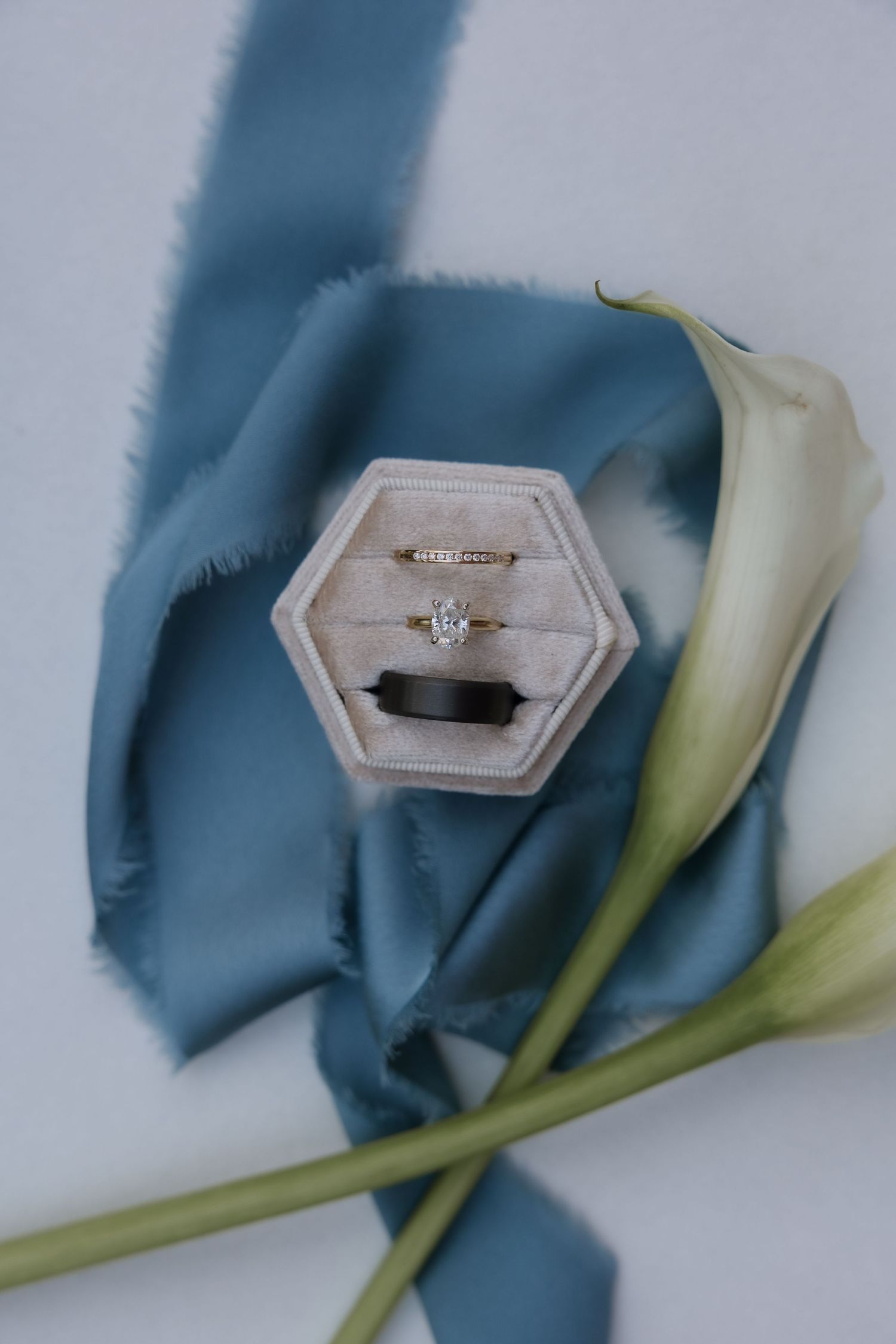 Elegant wedding rings in hexagonal velvet box with blue ribbon and white calla lily on light background.