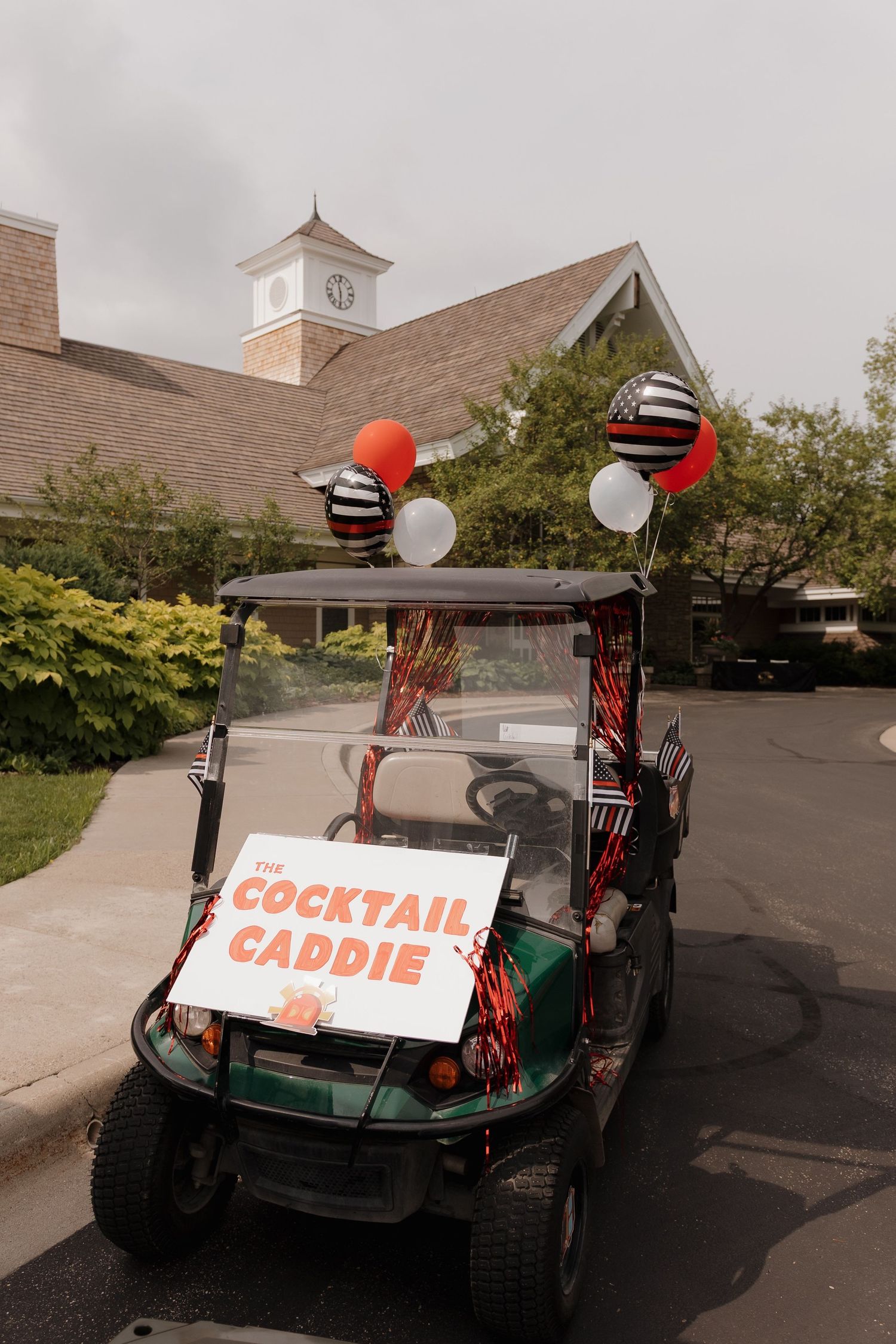 A decorated golf cart labeled The Cocktail Caddie with colorful balloons parked outside a clubhouse.