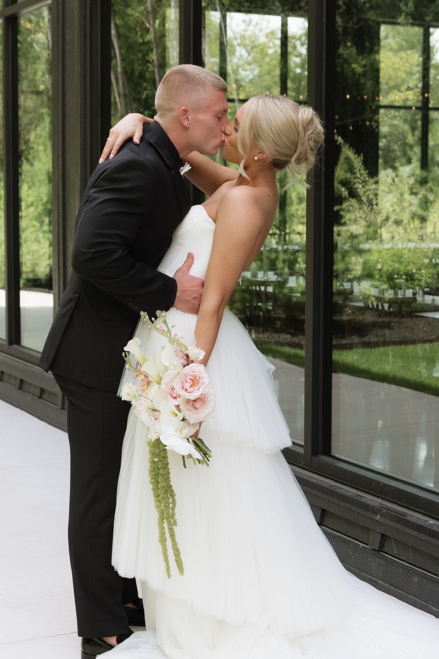 Bride and groom sharing a romantic kiss in a white wedding dress and black tuxedo, holding a pink and white floral bouquet.