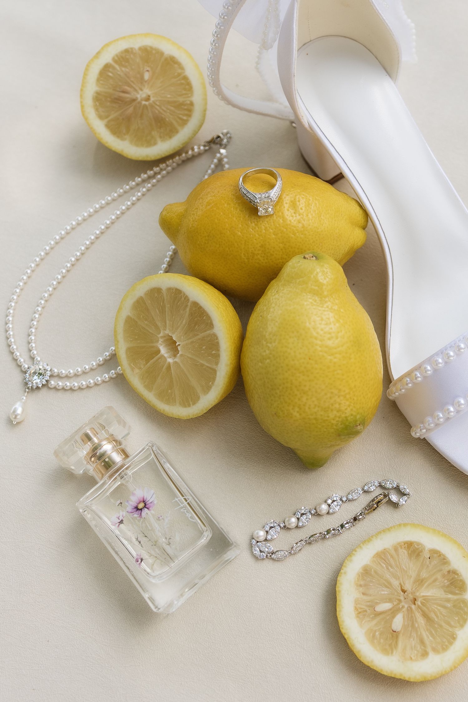 Bridal flat lay with lemons, white heels, diamond ring, pearl necklace, perfume bottle, and crystal bracelet on neutral background.