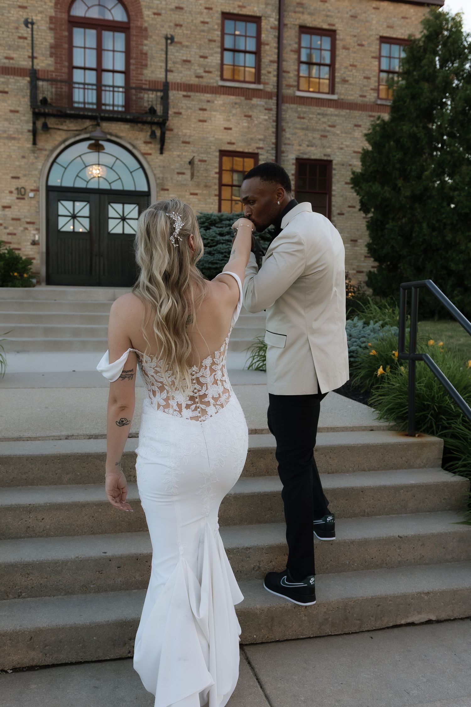 Groom in ivory blazer kissing bride's hand outside elegant stone venue, bride wearing backless white mermaid wedding gown.