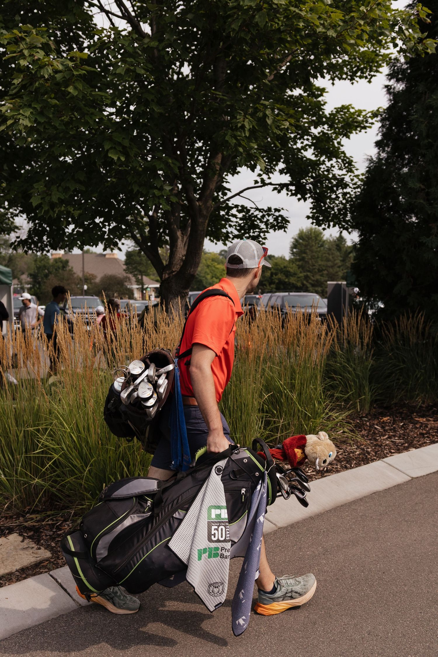Golfer in orange shirt carrying golf bag walking along path with ornamental grasses and trees in background.