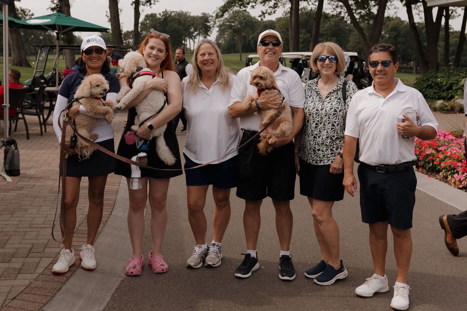 Group of people posing with small dogs on a golf course path on a sunny day.