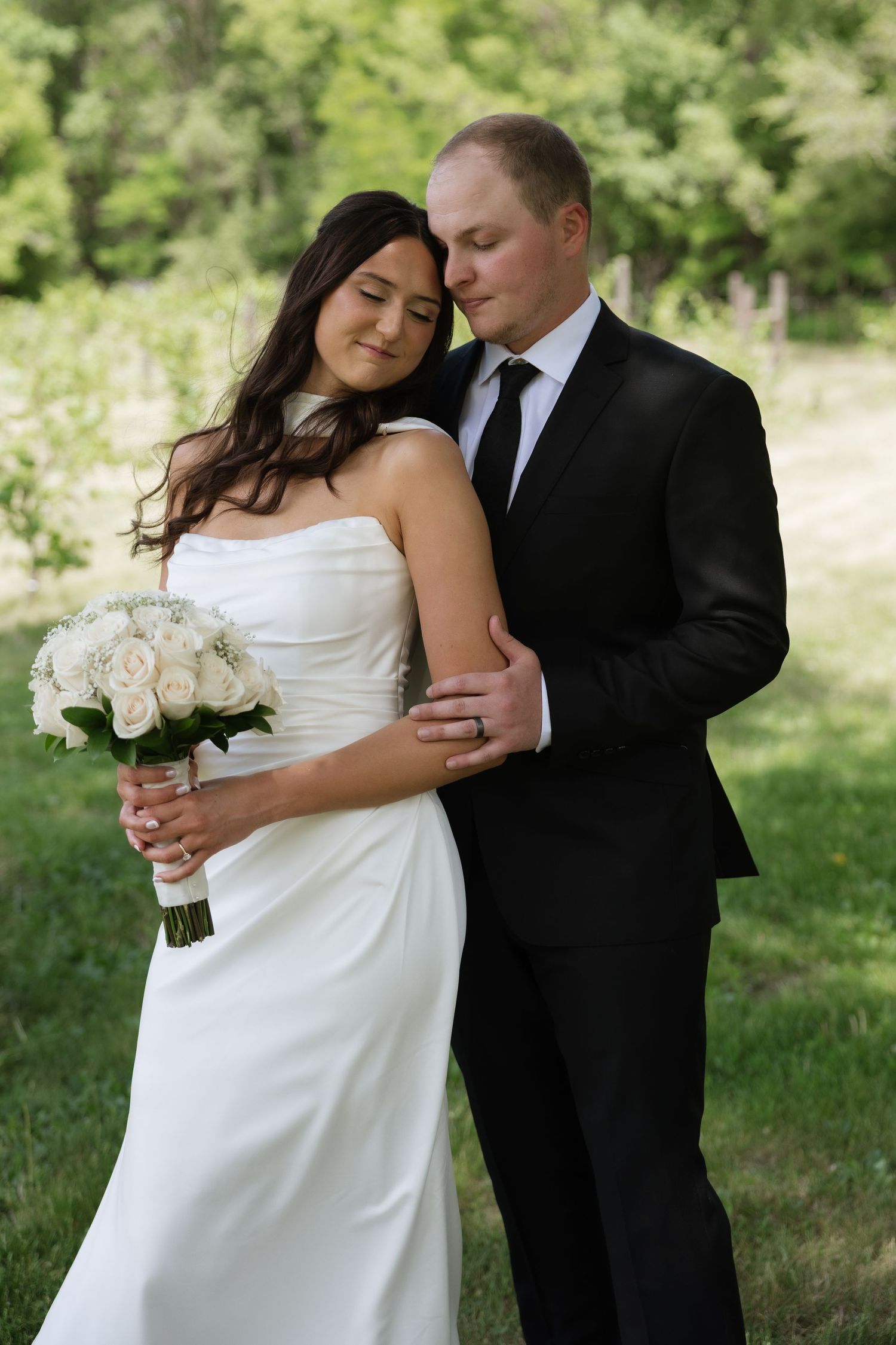 Groom in black suit tenderly embraces bride holding white rose bouquet in sunlit green garden.