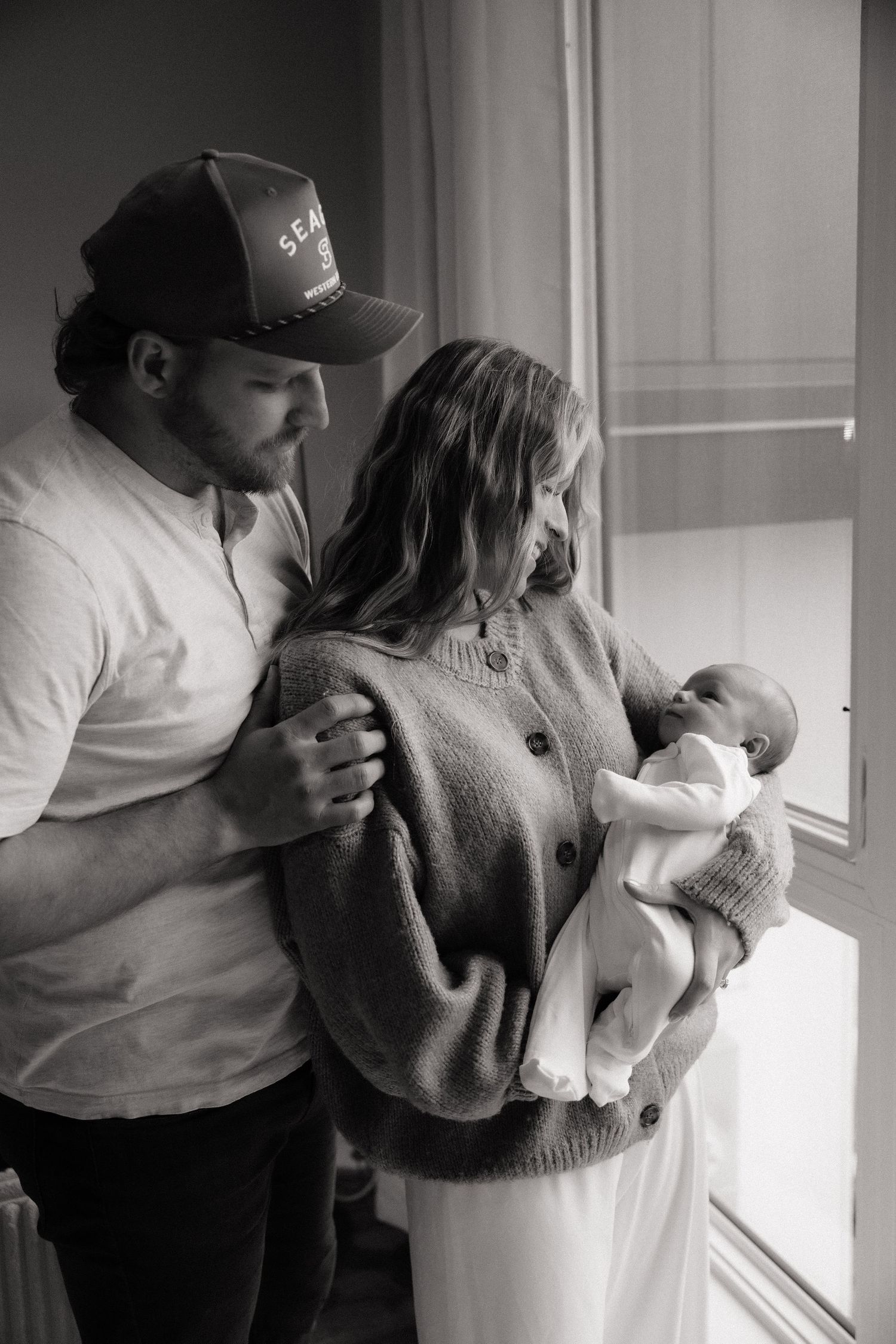 Black and white photo of parents lovingly gazing at their newborn baby near a bright window.
