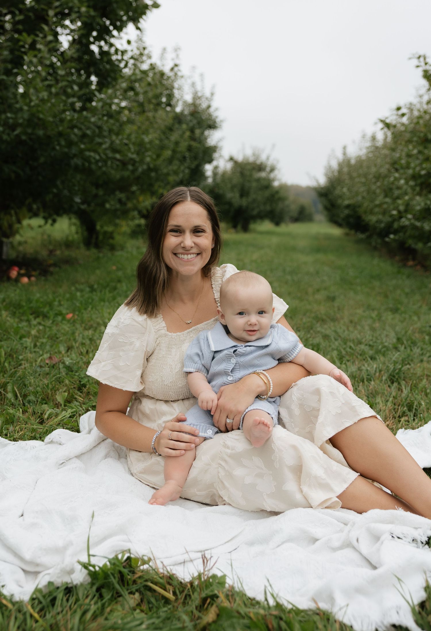 A smiling woman in a beige dress sits on a white blanket in an orchard holding a baby in a blue outfit.