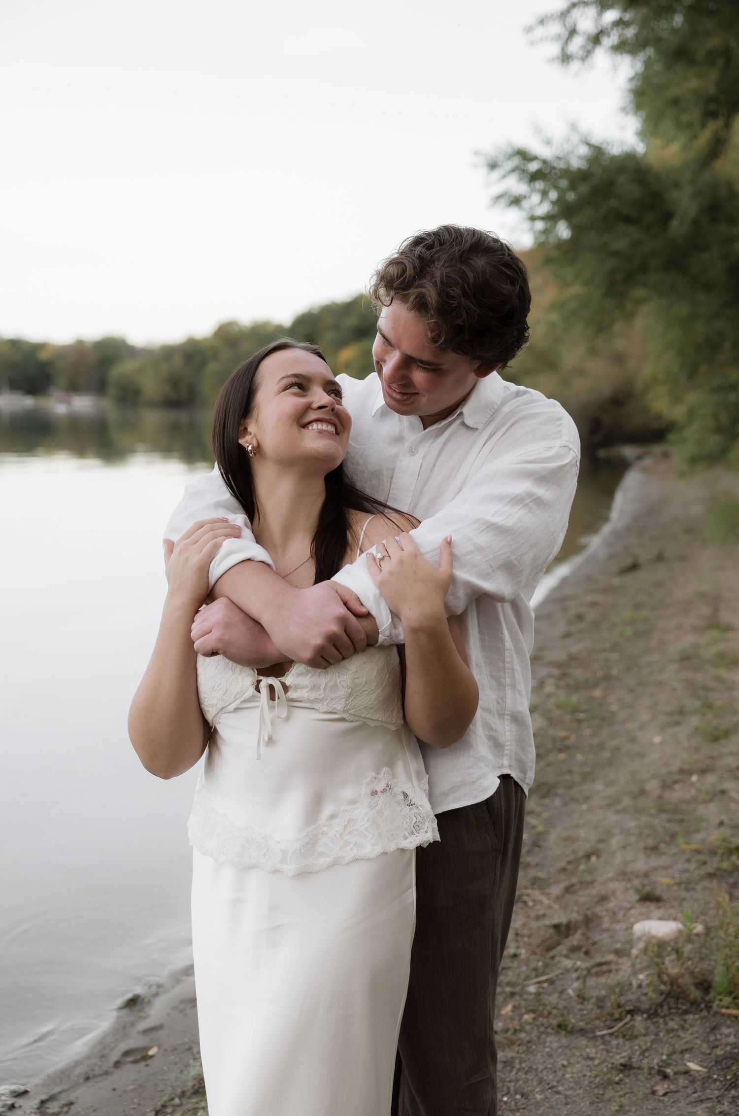 A couple embraces and smiles at each other on a riverside shore, the woman in white dress, man in white shirt.