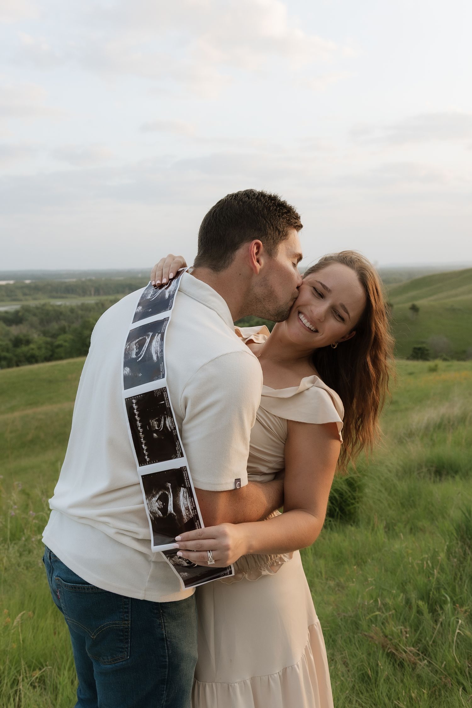 Couple embracing outdoors holding ultrasound photos, announcing pregnancy in a green meadow at sunset.