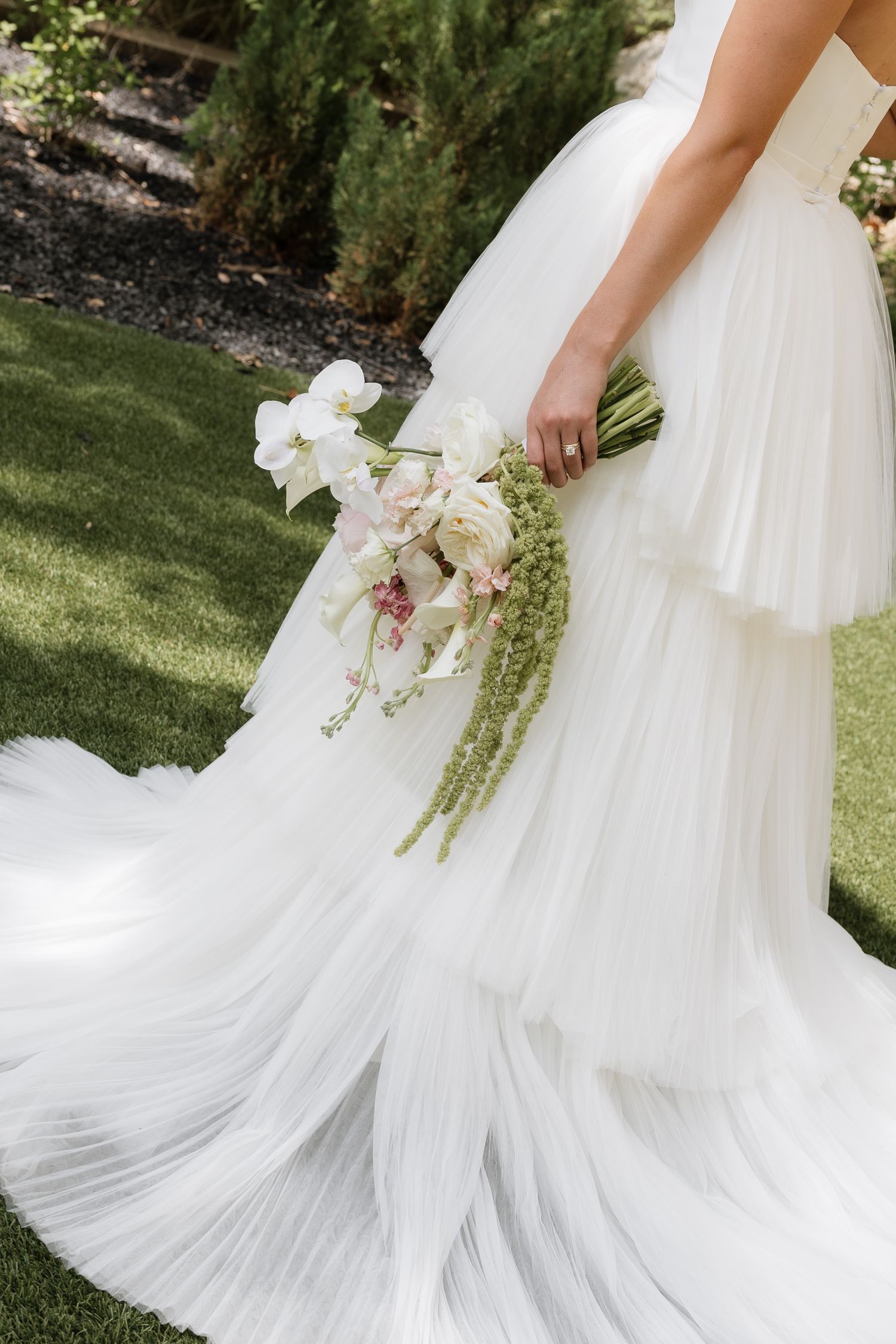 Bride in a layered white tulle wedding gown holding an elegant bouquet of white orchids and greenery outdoors.