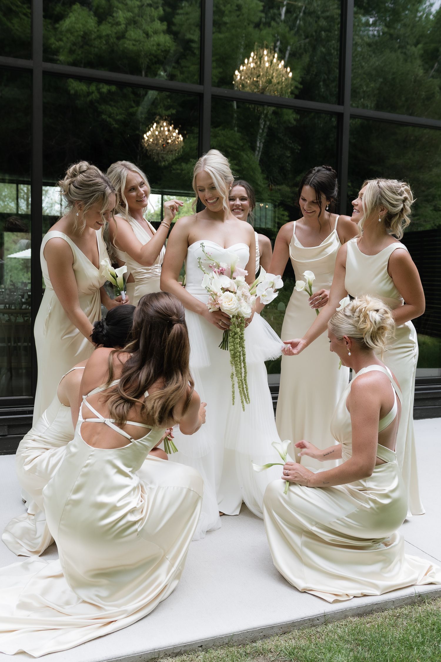 Bride in white strapless gown surrounded by bridesmaids in champagne satin dresses, admiring her bouquet outdoors.
