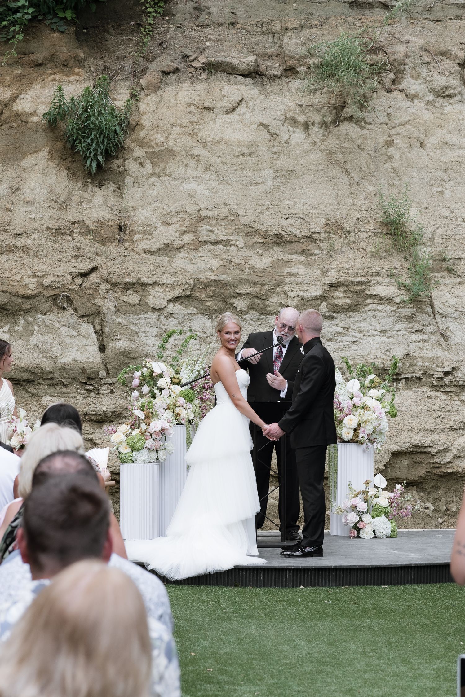 Bride and groom laughing during outdoor wedding ceremony against natural stone wall with white floral arrangements.