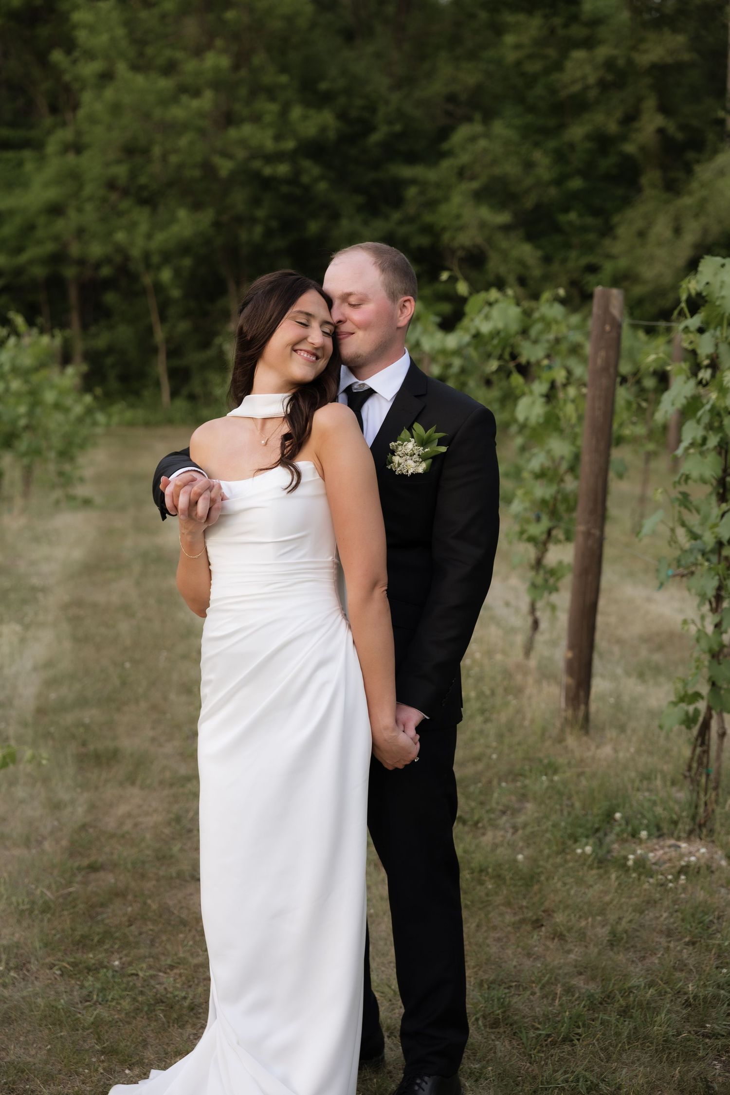 Bride and groom share a tender embrace in a vineyard, with the groom kissing her temple in a black suit and white gown.