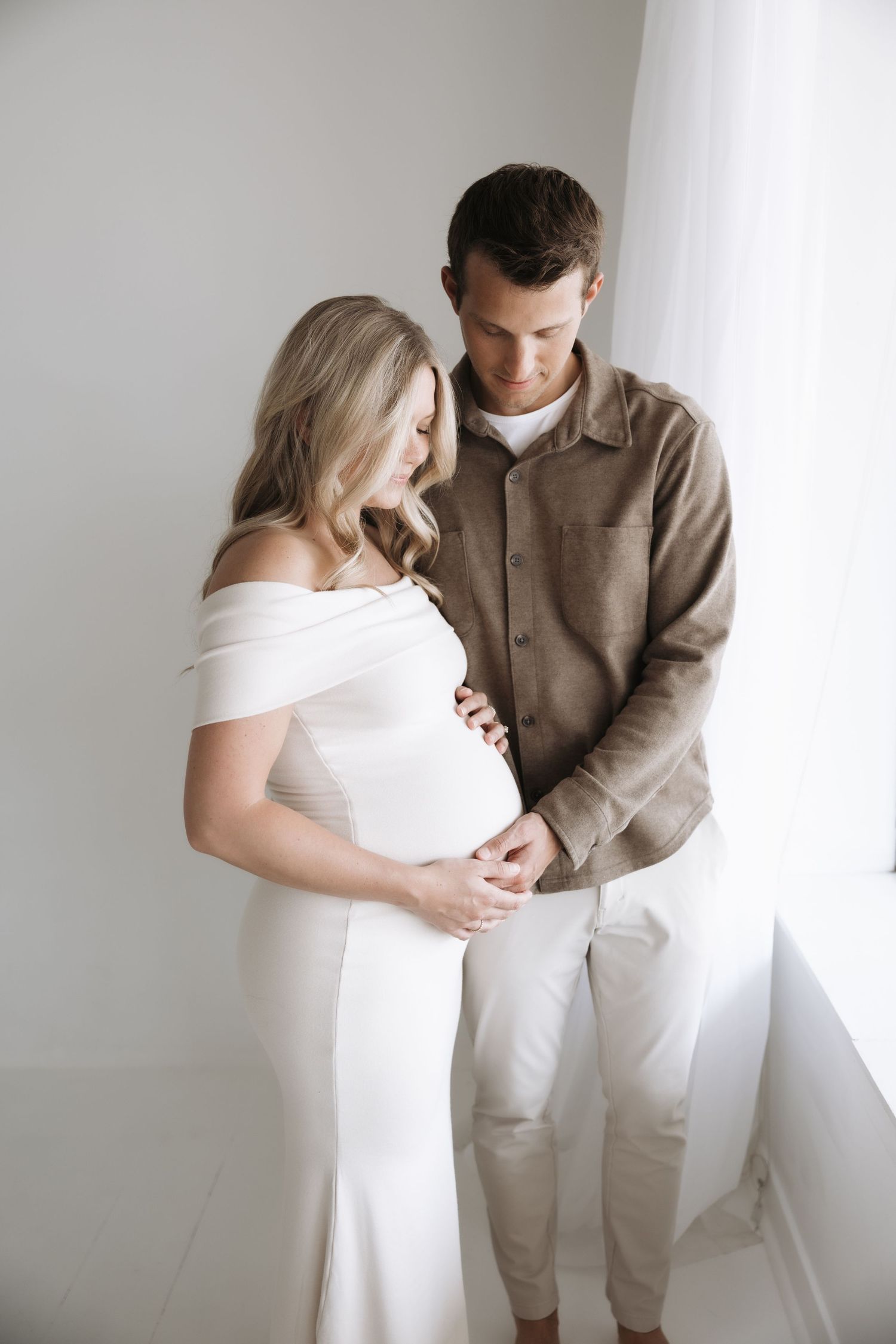 Couple during maternity shoot holding hands over pregnant belly near bright window.