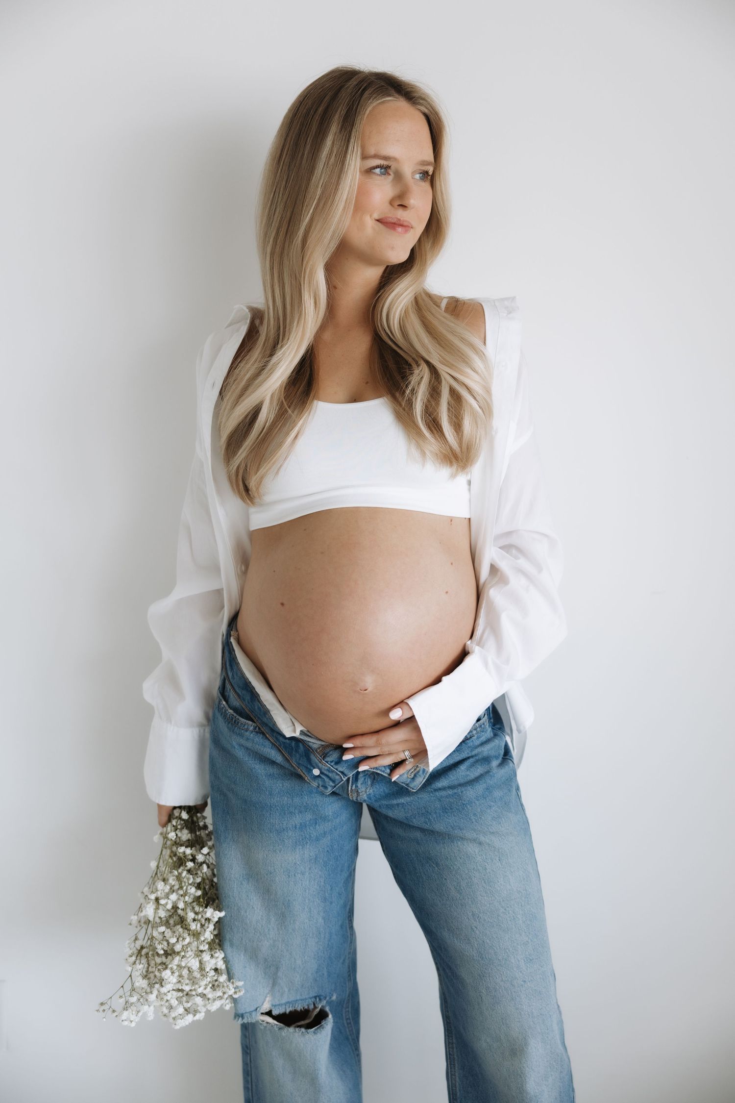 Smiling pregnant blonde woman in white open shirt, crop top, and ripped jeans holding baby's breath bouquet.