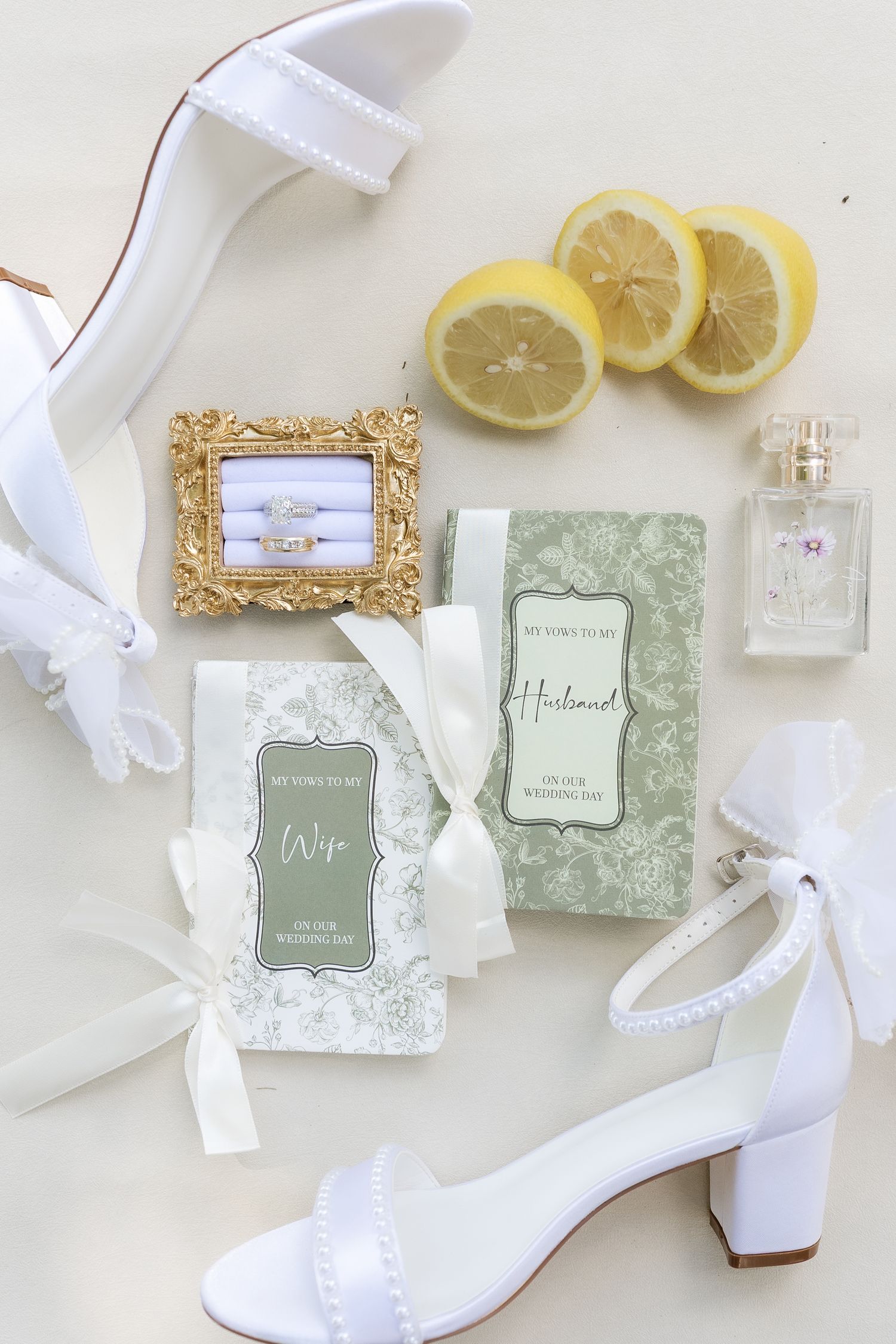 Flat lay of wedding details including white heels, rings, vows books, perfume, and lemons.