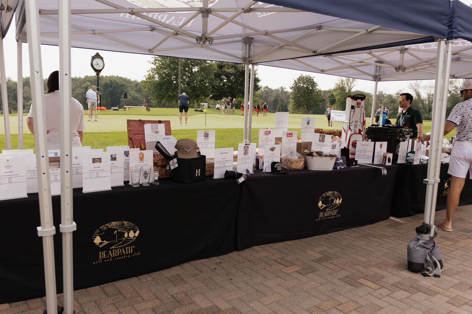 Outdoor vendor booth under a tent at a golf course event, displaying various products on a black-draped table.