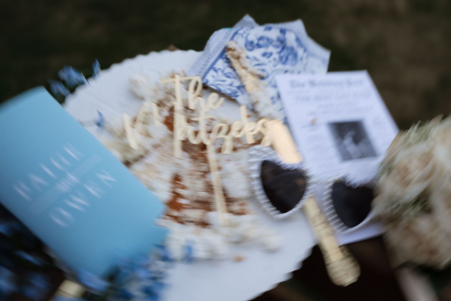 Blurred close-up of wedding flat lay featuring blue program, heart sunglasses, and decorative cake topper.