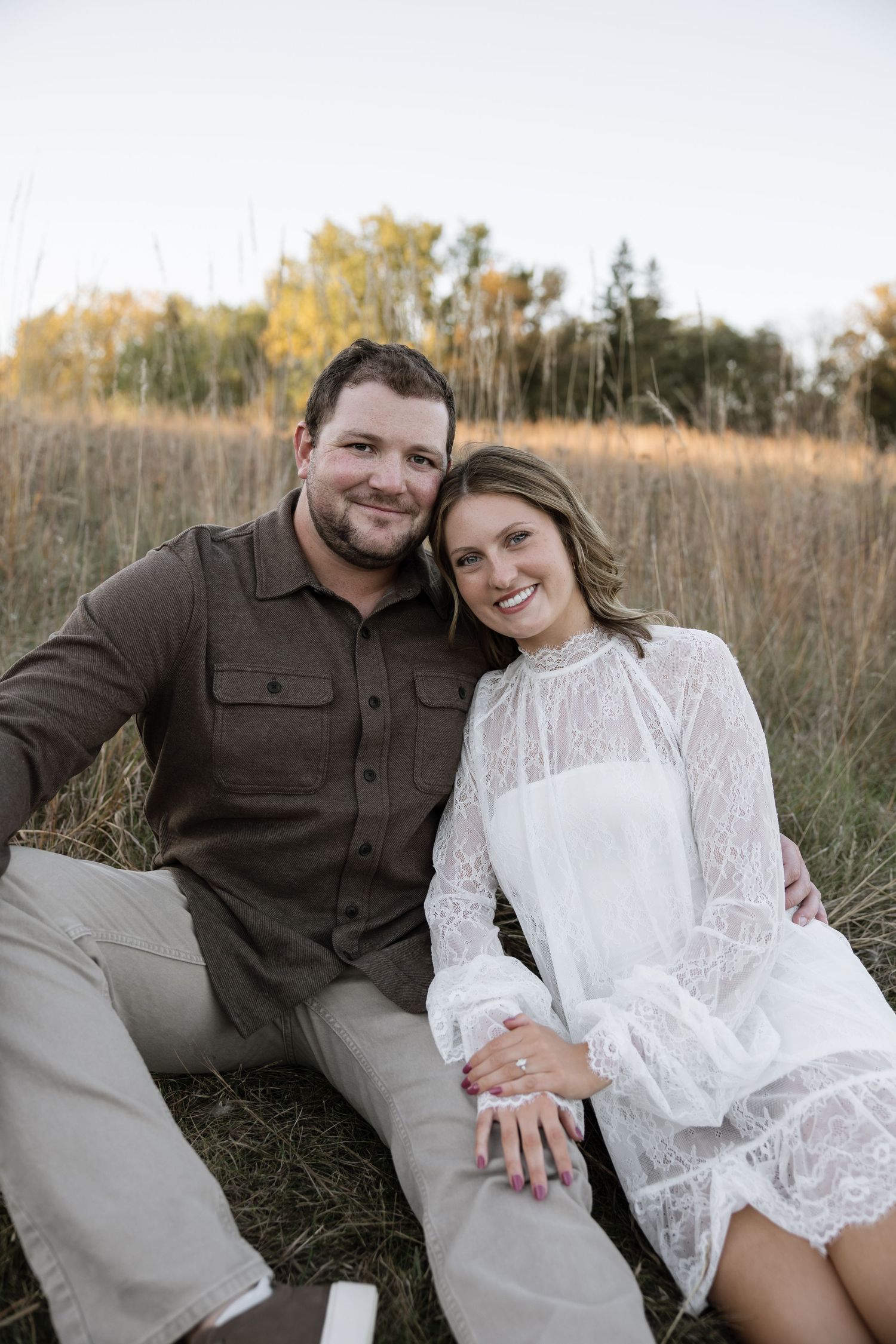 Couple sitting in golden field, woman in white lace dress showing engagement ring, man in brown shirt smiling.