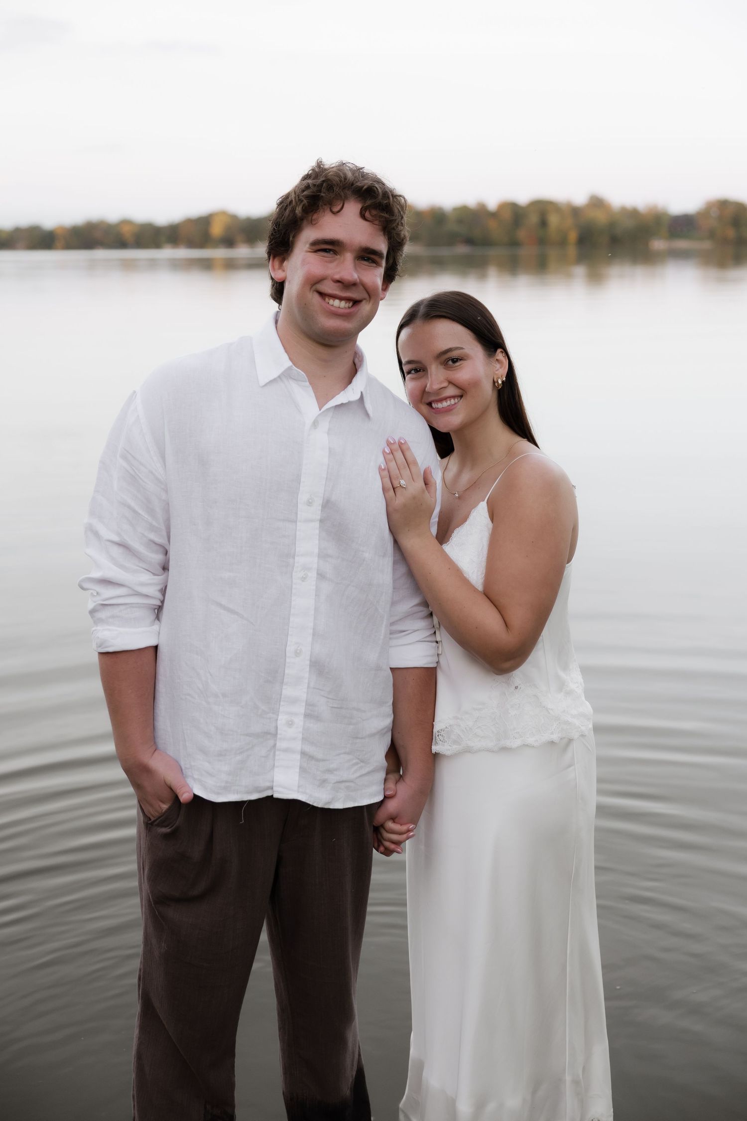 Happy engaged couple standing in lake water, bride showing off diamond ring to camera at sunset.