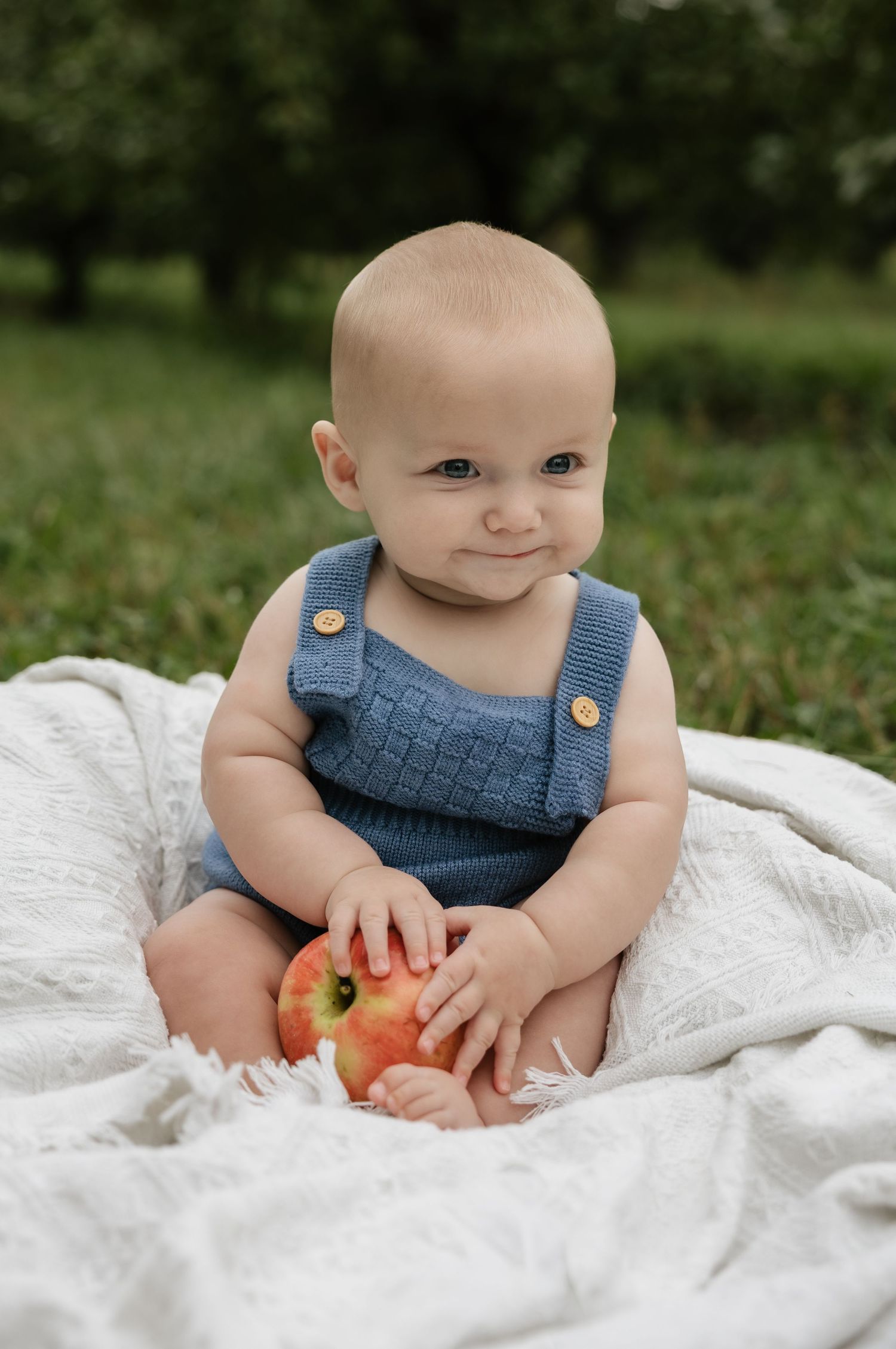 Baby in blue knit overalls sitting on white blanket outdoors, smiling while holding a red apple in a green orchard.