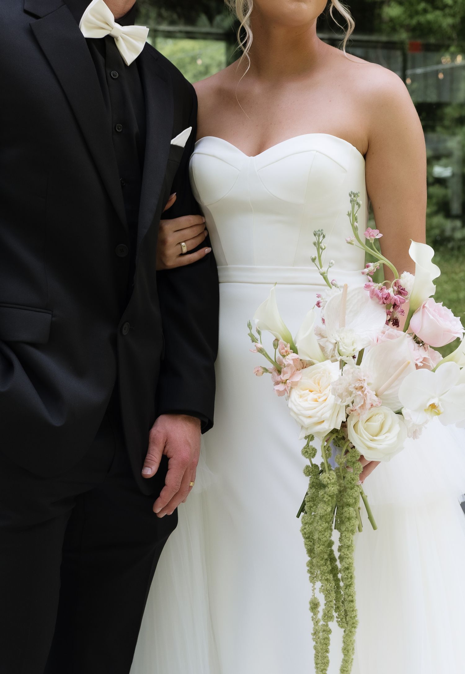 Bride in strapless white gown holds pink and white bouquet beside groom in black tuxedo with bow tie.