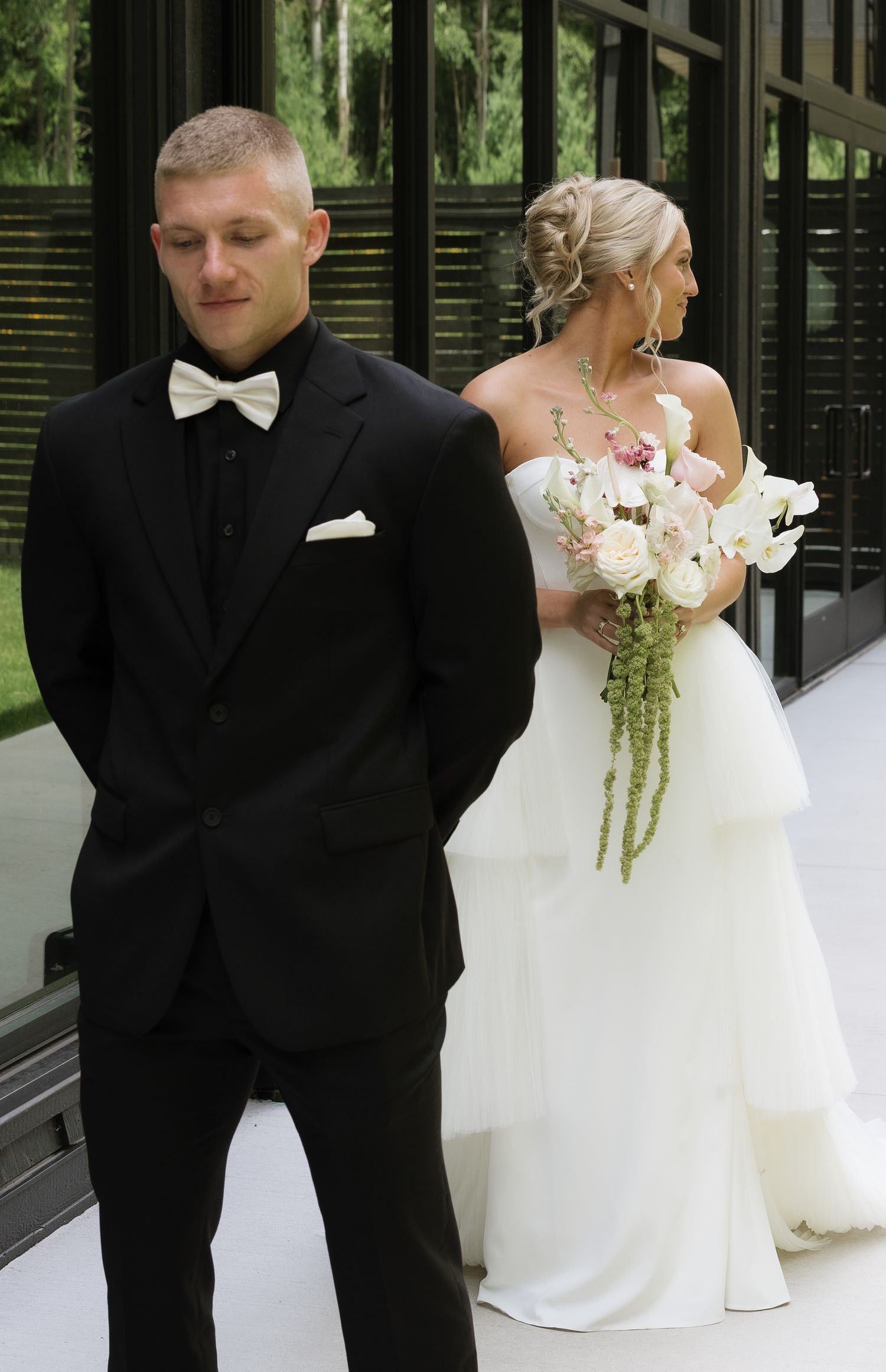 Emotional groom in black tuxedo standing back-to-back with bride in strapless white gown before first look.