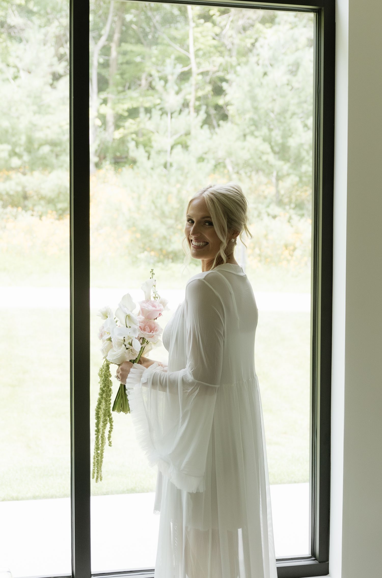 Smiling bride in white sheer gown holding floral bouquet standing by large window with green trees in background.