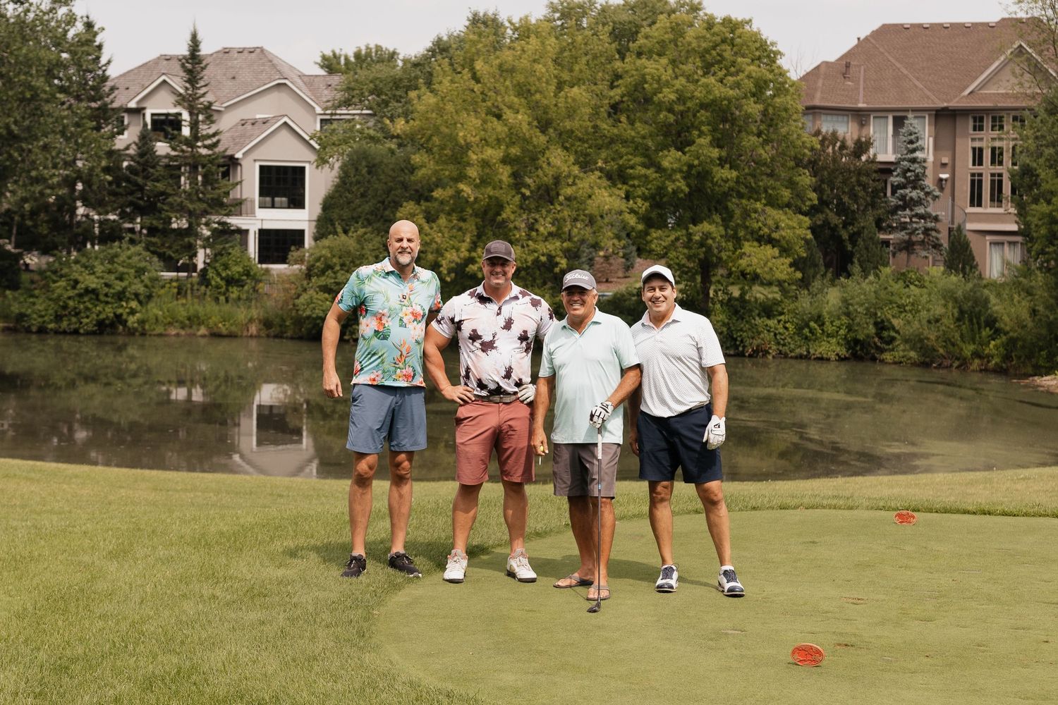 Four golfers posing together on a lush green golf course with a pond and houses in the background.
