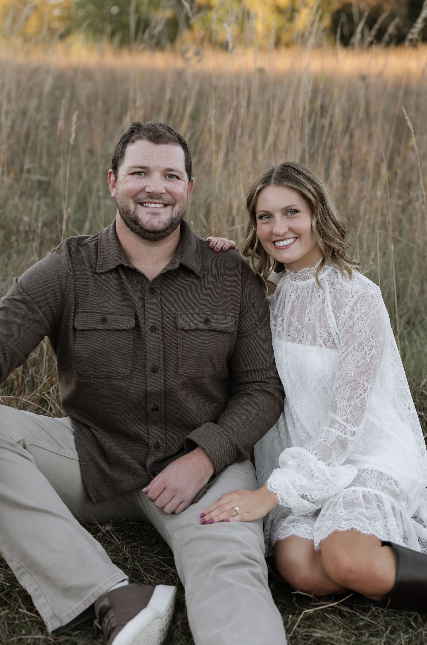 A smiling couple poses outdoors in a field, man in brown shirt and woman in white lace dress, likely an engagement photo.