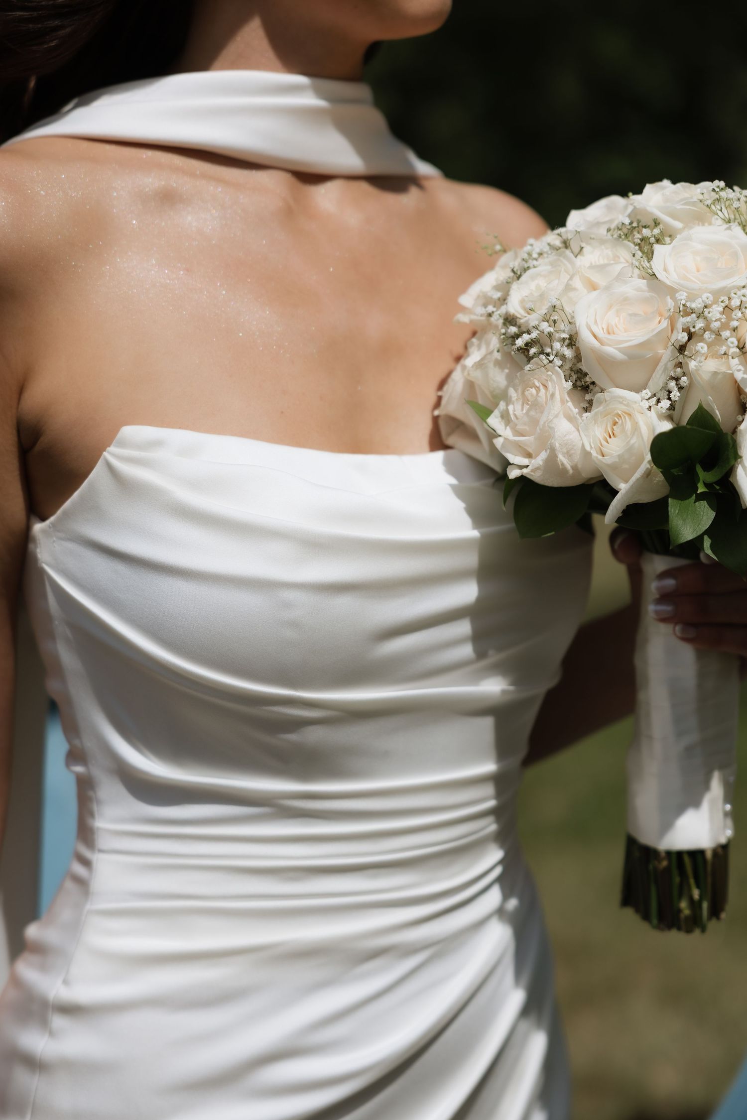 Close-up of bride in elegant strapless white satin gown holding white rose and baby's breath bouquet.