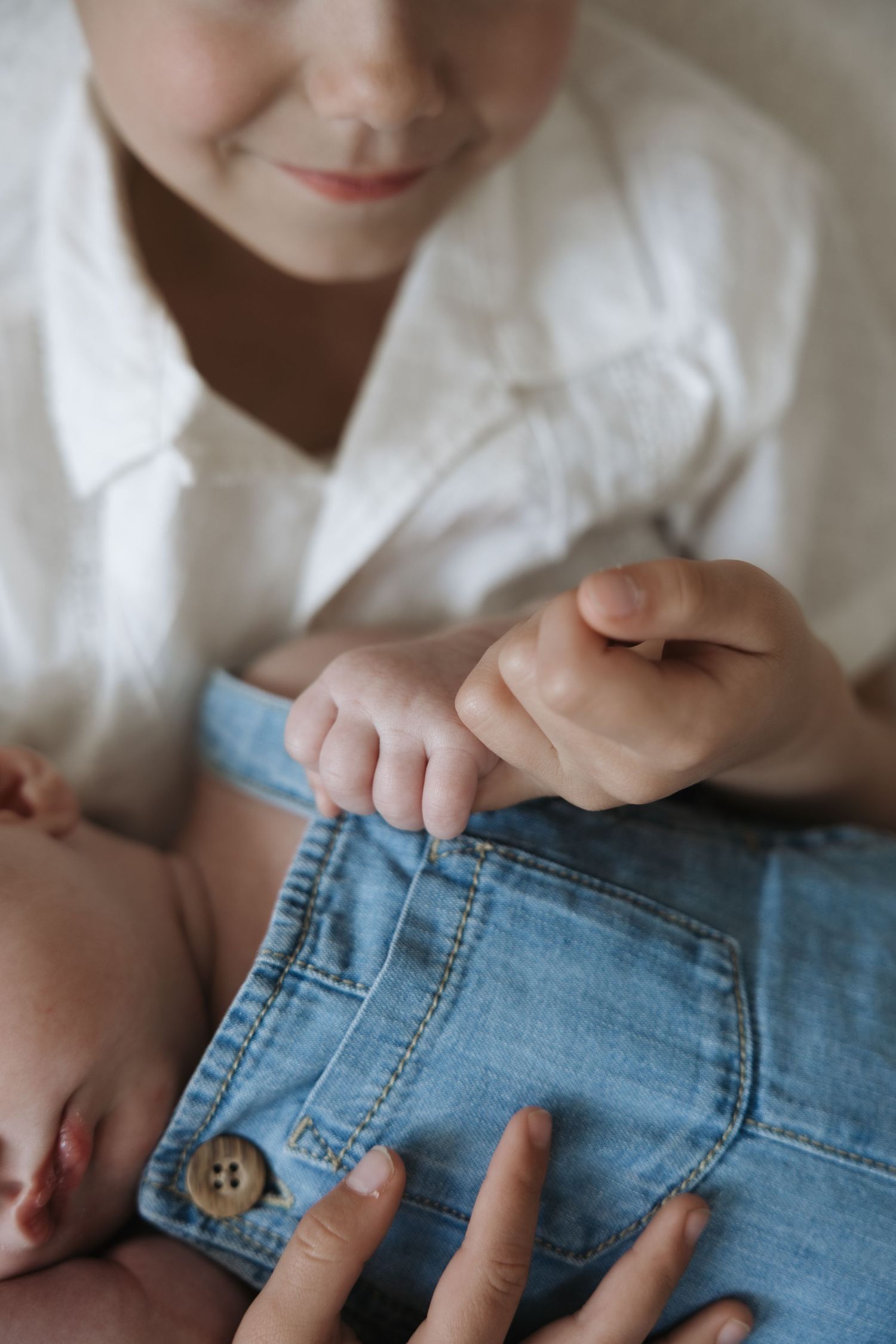 Close-up of older child holding newborn baby's tiny hand while baby wears a blue denim romper.