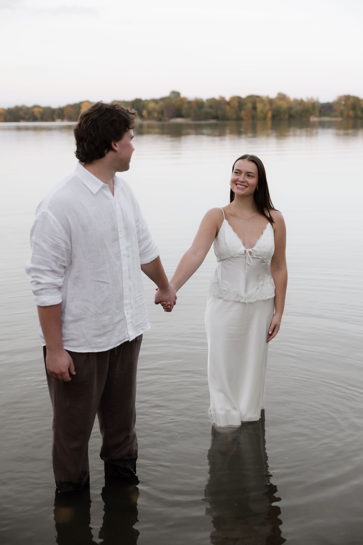 Couple holding hands while standing in calm lake water, woman in white dress smiling at man in white linen shirt.