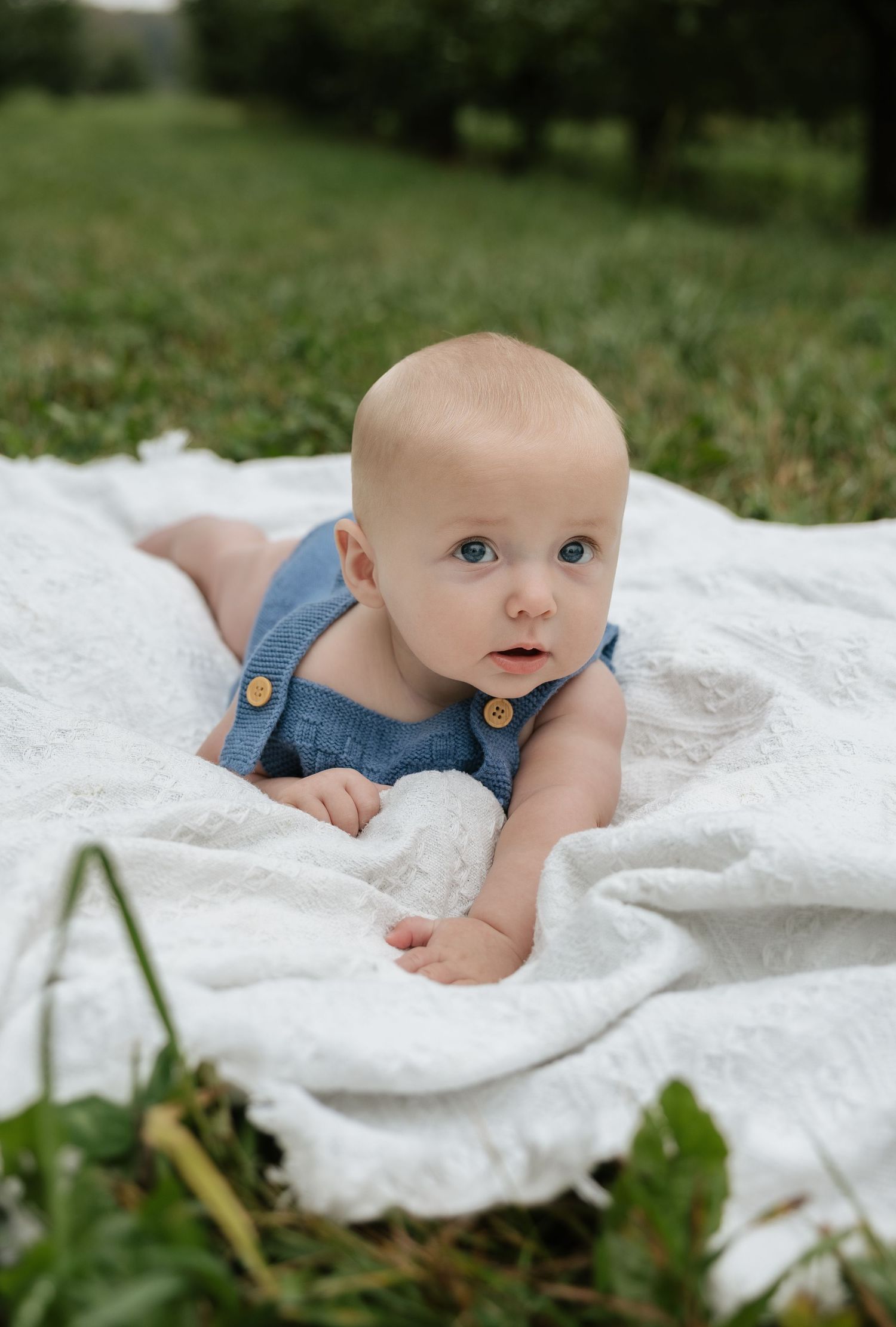 Baby in blue overalls doing tummy time on white blanket in green grass outdoors.