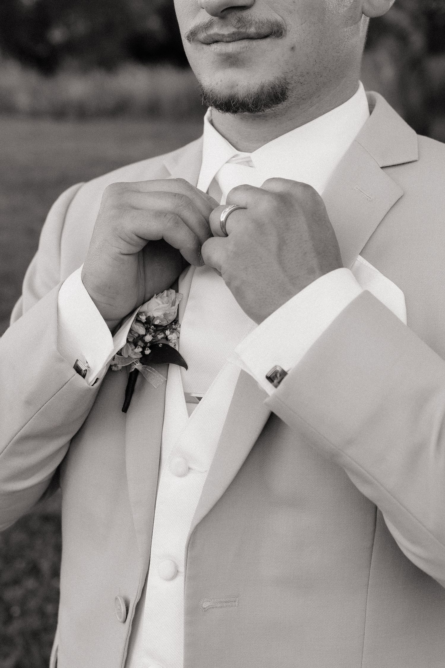Black and white photo of a groom in a light suit adjusting his tie, wearing a boutonniere and ring.