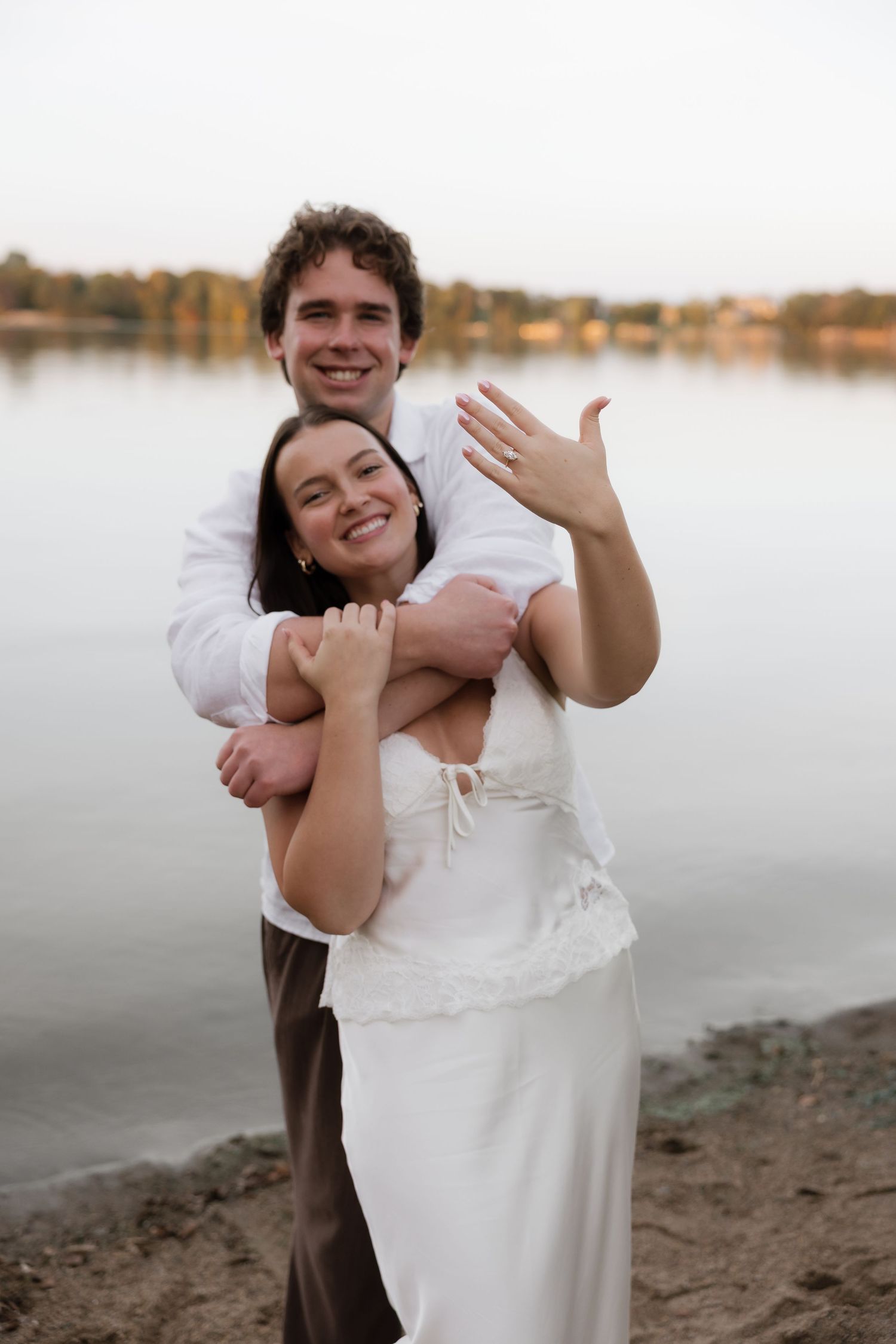 Engaged couple embracing on lakeside beach, woman showing off ring, both smiling joyfully in golden hour light.