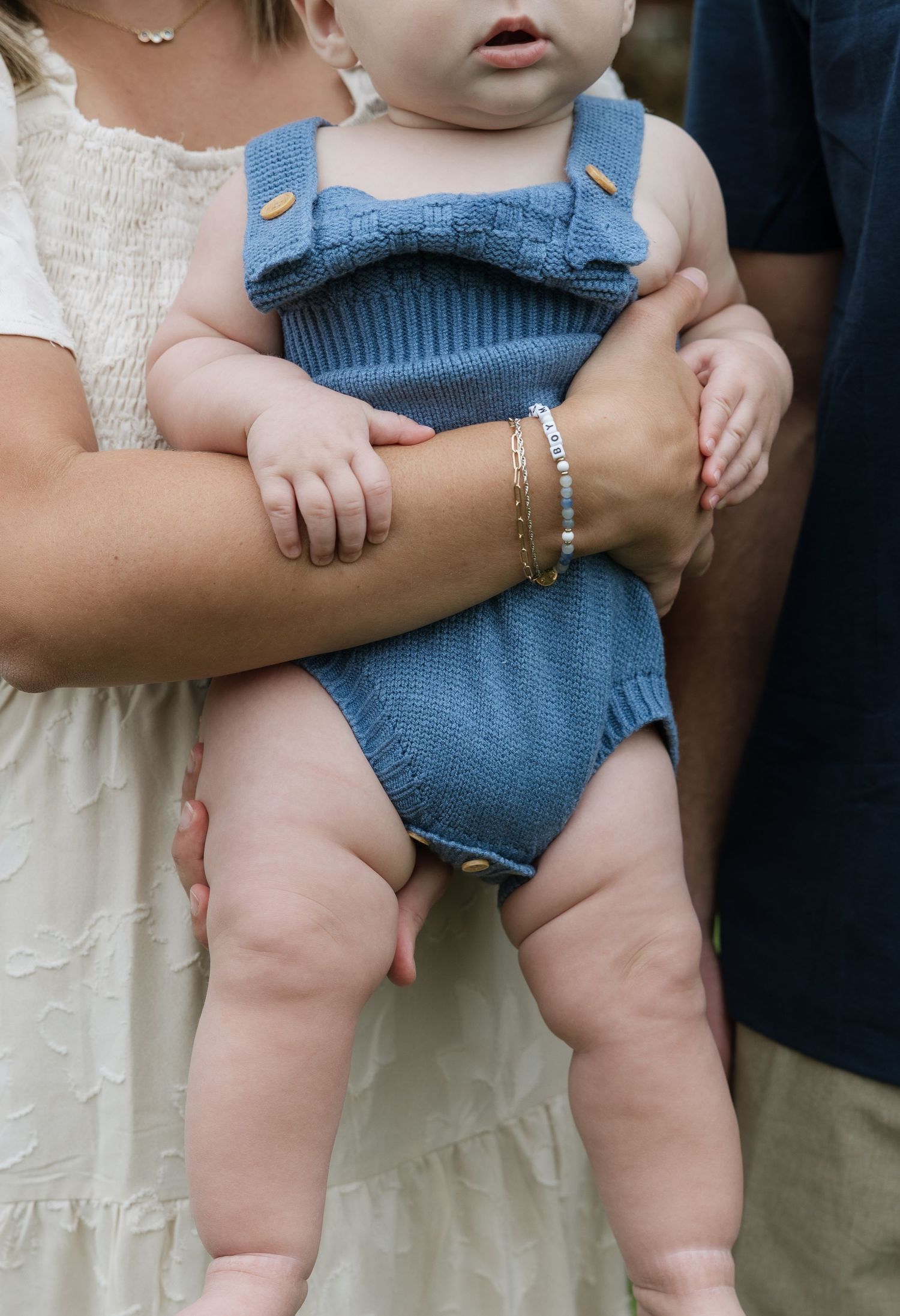 Baby in blue knit romper held by parents, showing chubby legs and mother's beaded bracelet.