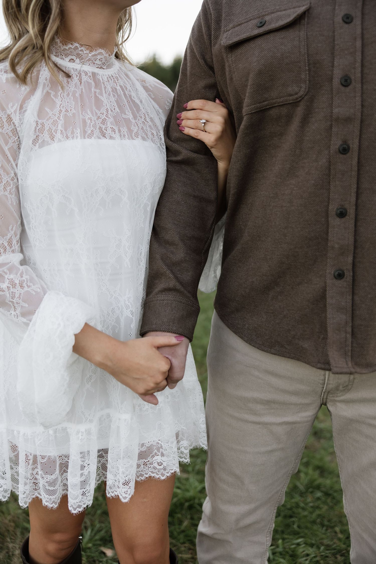 Couple holding hands, woman in white lace dress with ring visible, man in brown shirt and khaki pants.